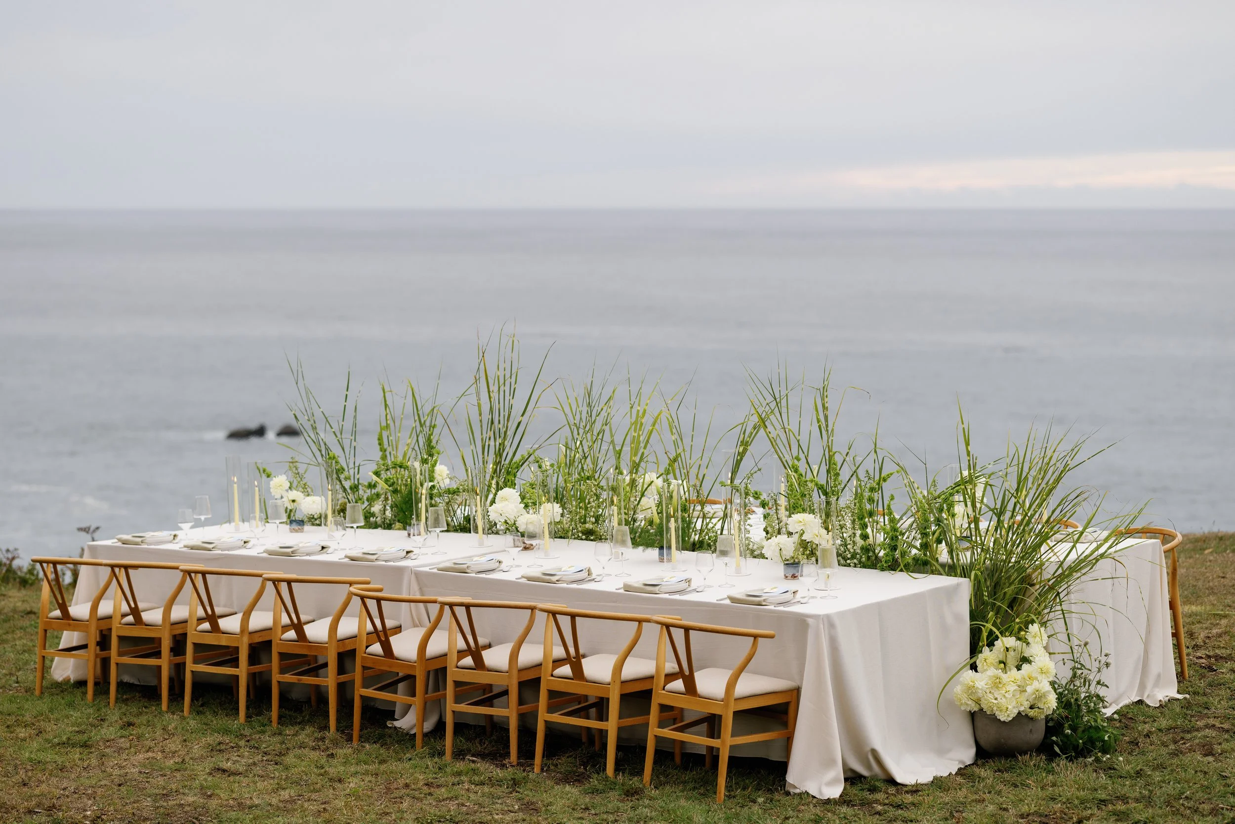 Long rectangular outdoor dining table set with white tablecloth, white plates, silverware, glasses, and tall candles, positioned on grassy area next to a beach with ocean and cloudy sky in the background, decorated with white flowers and greenery.