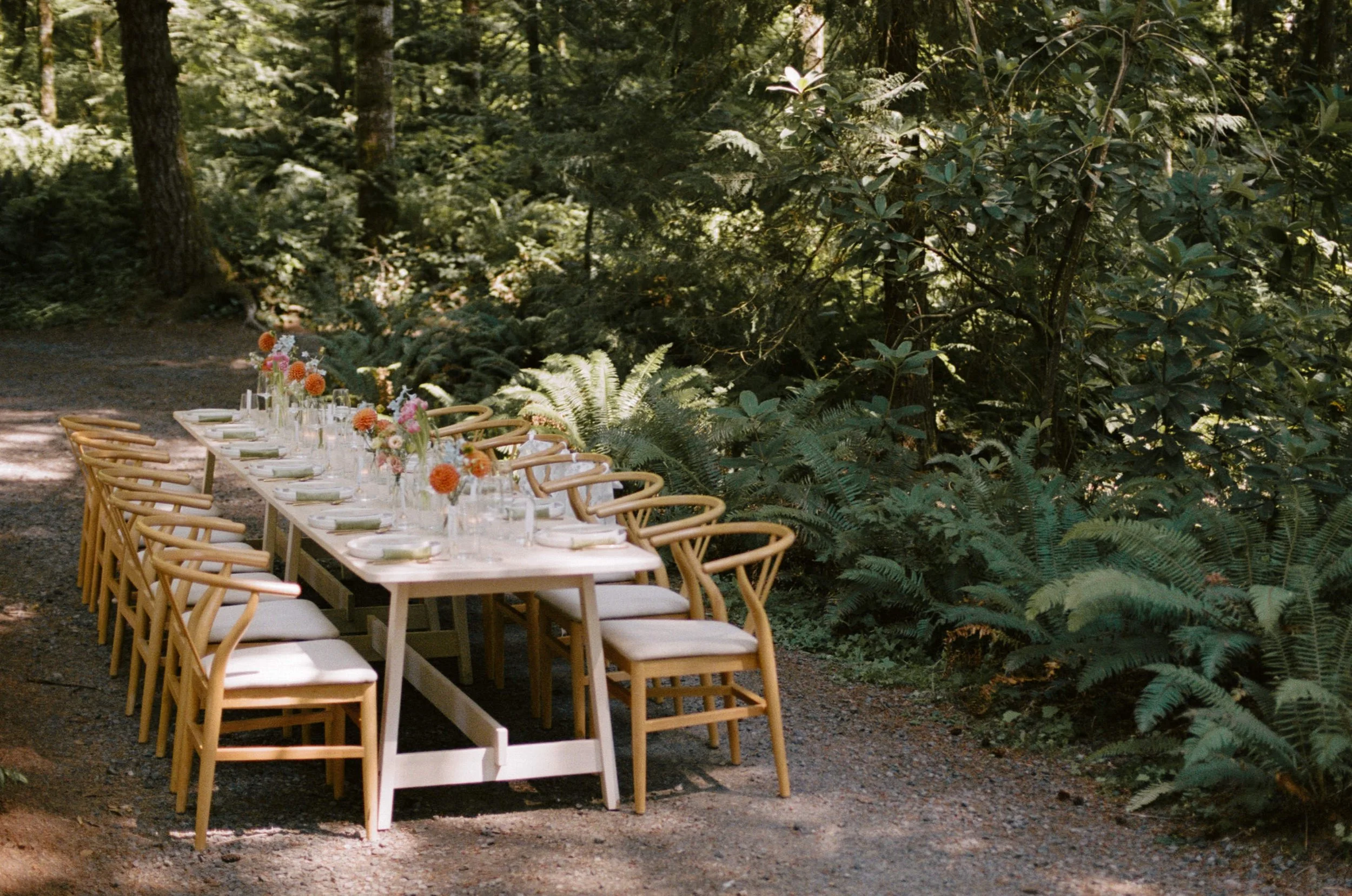 A long wooden table set for an outdoor meal, decorated with colorful flowers in vases, surrounded by wooden chairs, in a forested setting with trees and lush greenery.
