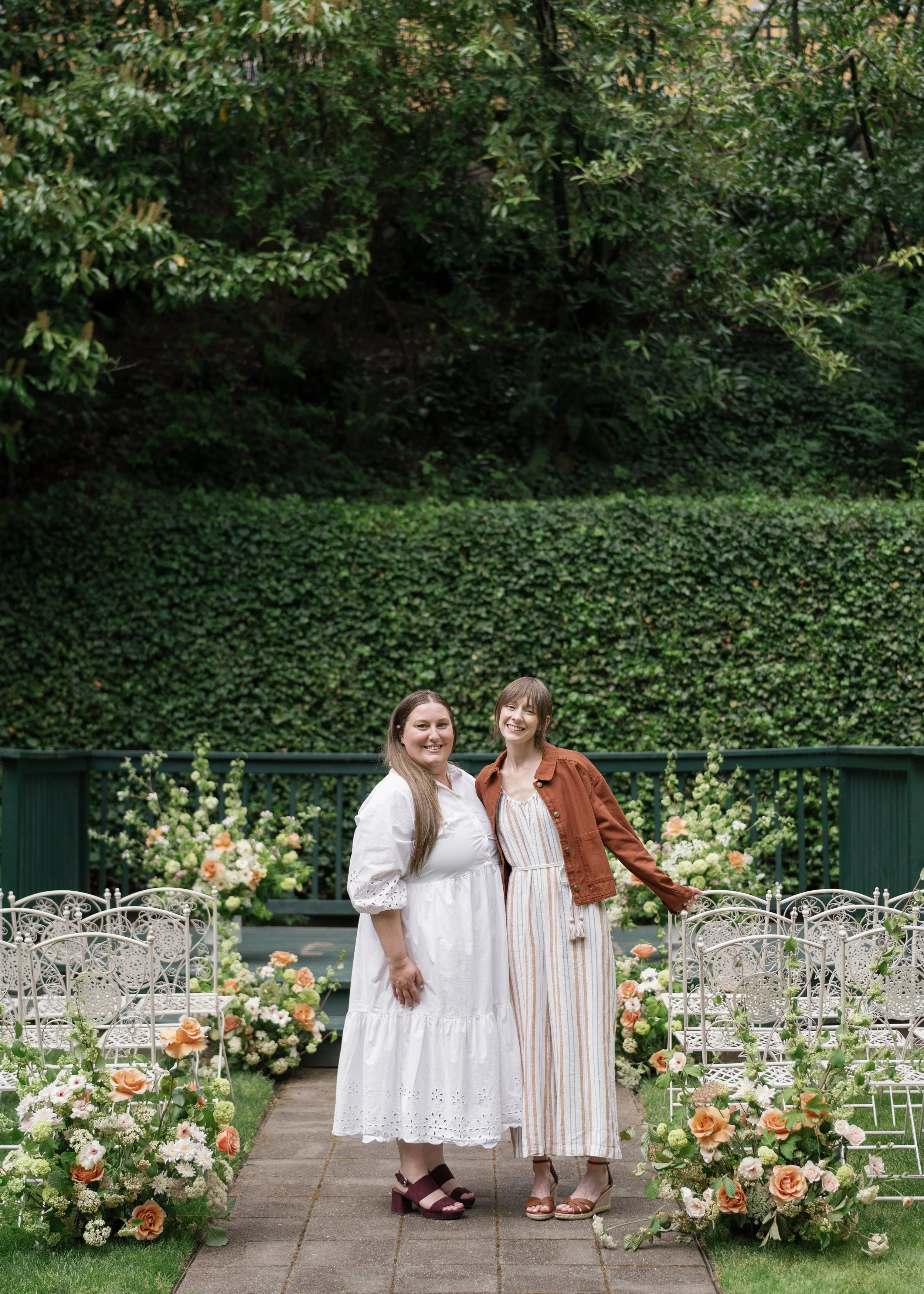 Two women standing together in a garden with flowers and greenery, smiling at the camera.
