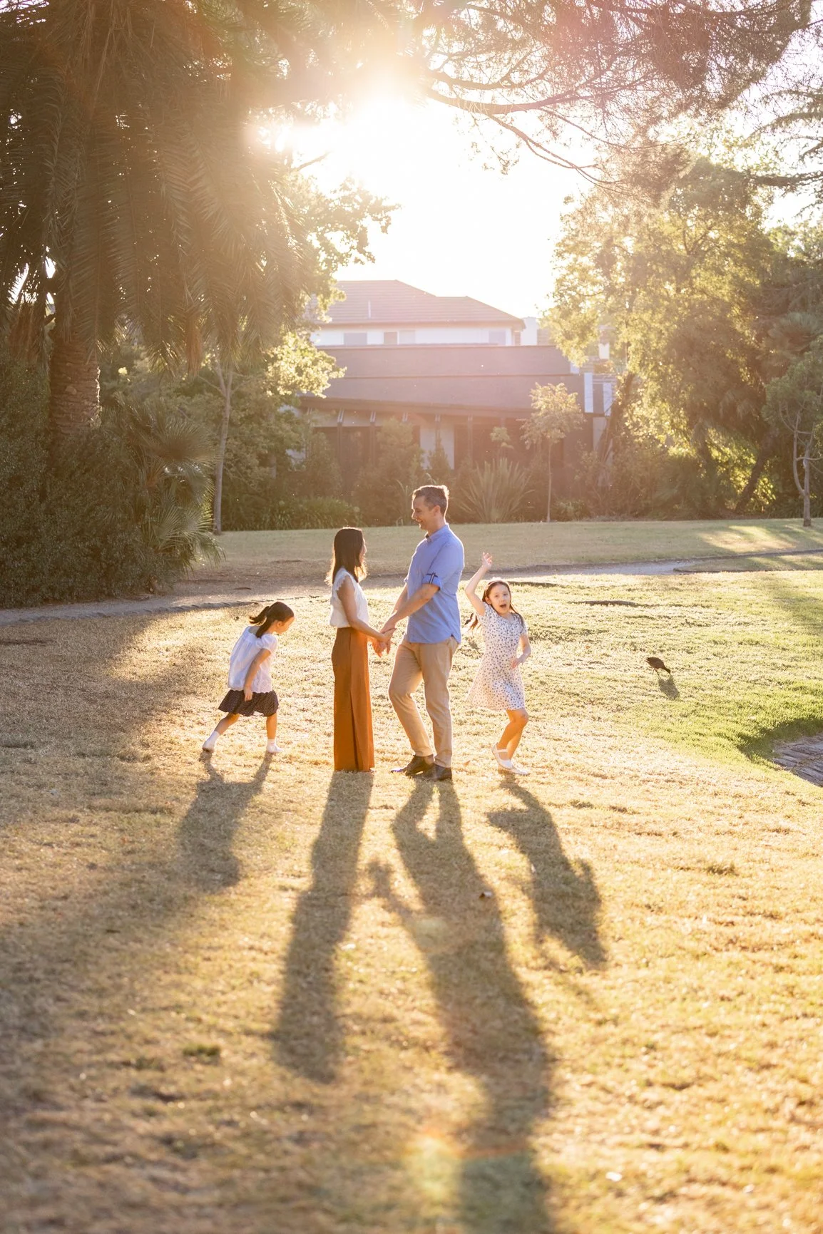 Family photo of four enjoying time outdoors in a sunny park, with parents holding hands and two children playing nearby, surrounded by trees and grass at sunset in Melbourne.