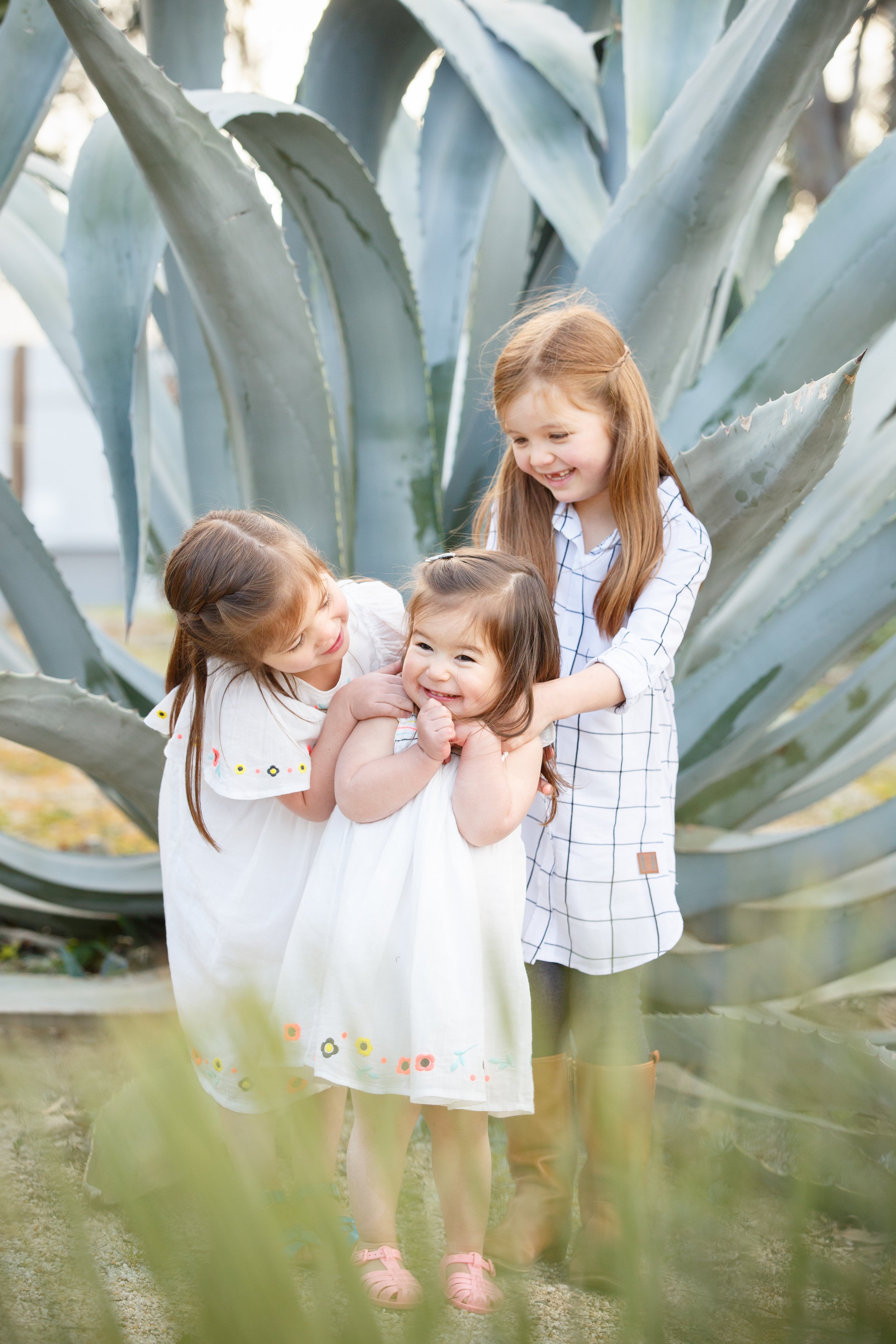 Family photo of three young girls smiling and standing together in front of large agave plants, wearing casual dresses in Melbourne.