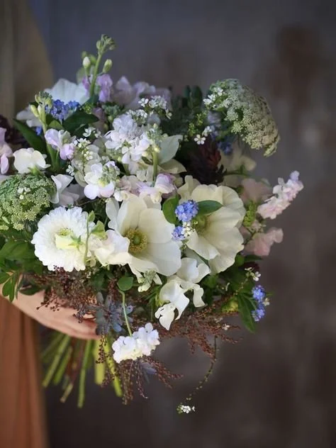 Hand holding a bouquet of white, purple, and blue flowers with greenery.