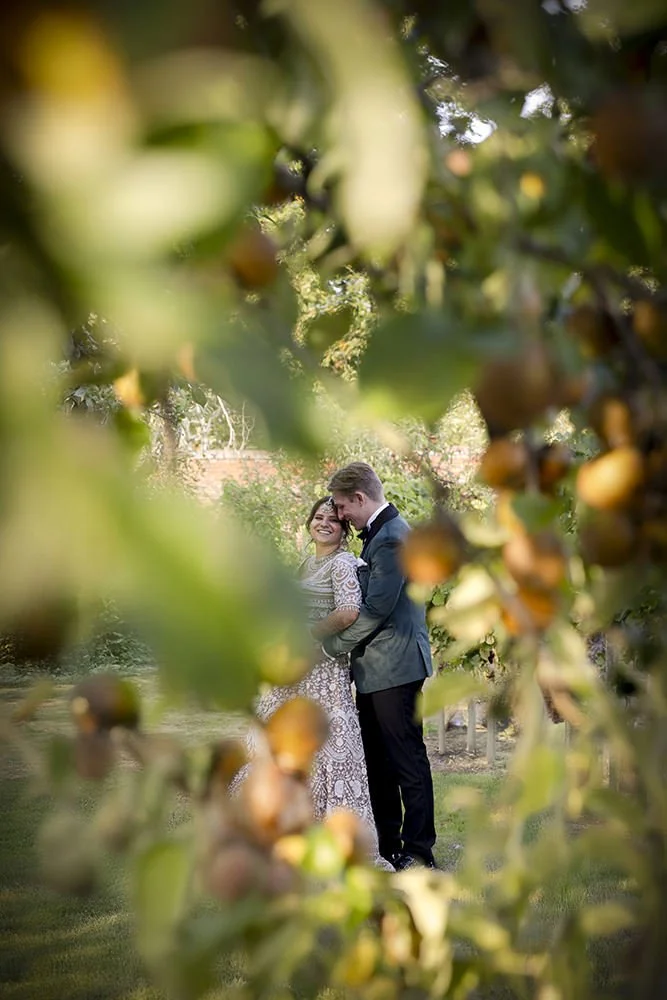 A couple in wedding attire smiling and embracing outdoors, framed by blurred green and brown foliage.