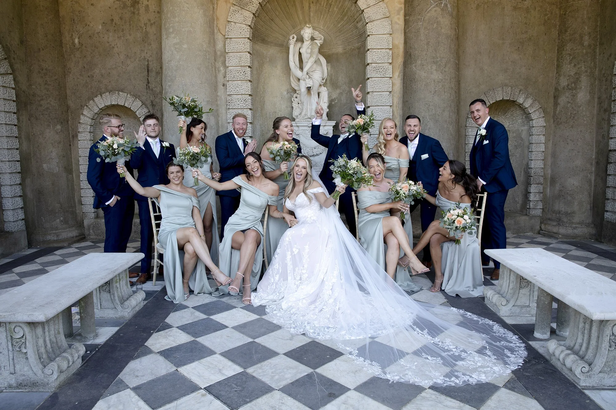 A wedding party poses joyfully in formal attire, with bridesmaids in matching dresses and groomsmen in suits, around a seated bride in a white gown, set against a stone architectural backdrop with a statue and checkered floor.