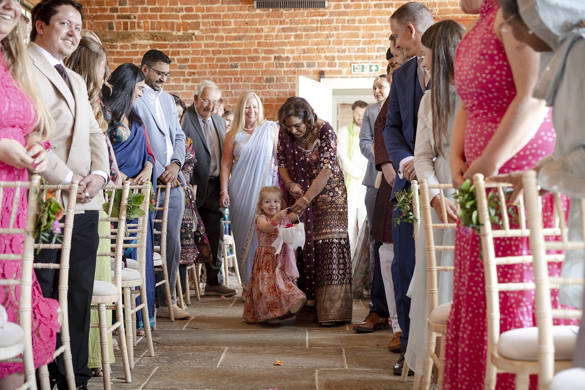 A young girl in a floral dress, holding a bag, is receiving a gift from a woman in traditional Indian attire, surrounded by guests in formal and traditional outfits at a wedding ceremony.