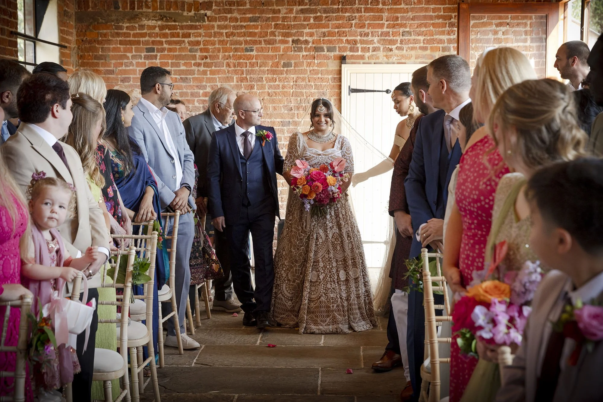 Bride in a wedding dress holding a colorful bouquet, standing between her parents, formally dressed, during a wedding ceremony indoors with guests seated on either side.