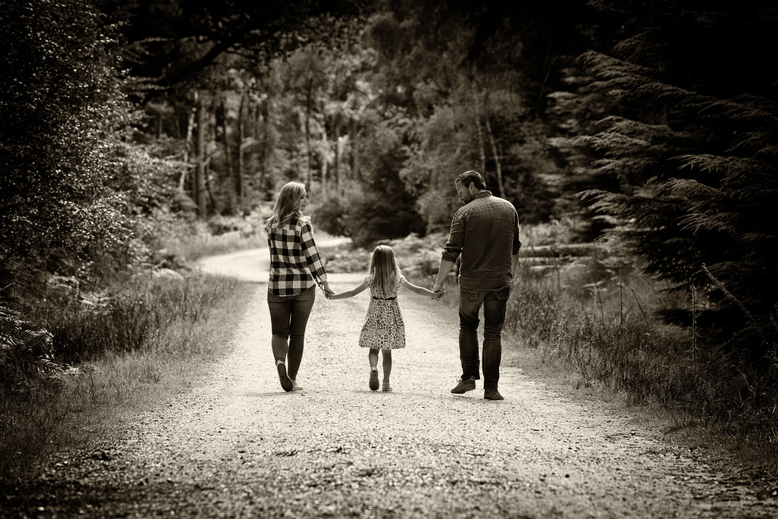 A family of three, a man, a woman, and a girl, walking hand-in-hand on a dirt path through a wooded area.