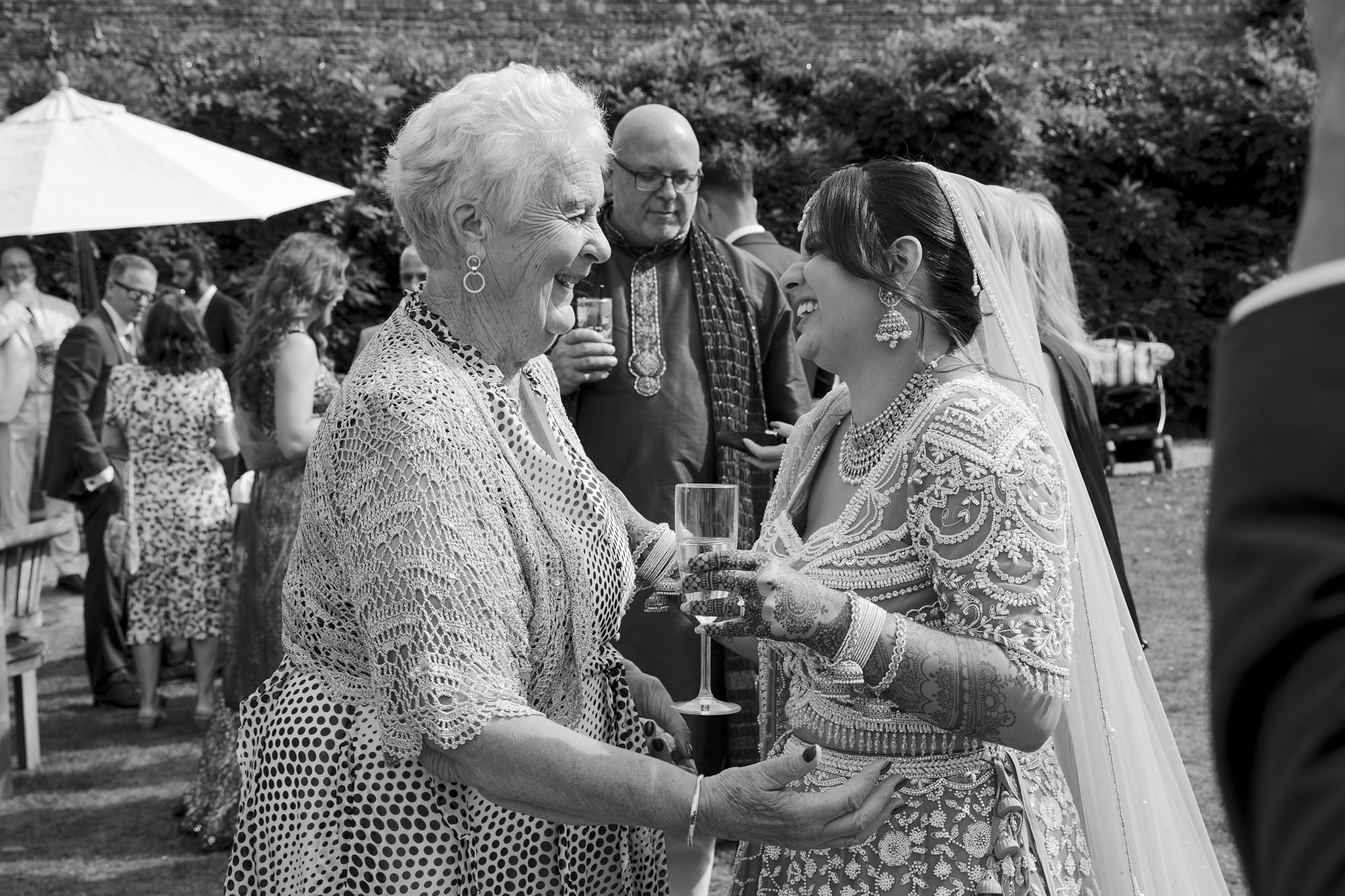 A joyful bride and an older woman share a happy moment at a wedding, holding glasses of champagne and smiling at each other. The bride is dressed in ornate traditional attire and jewelry, while the woman wears a polka dot dress and a crochet jacket. 