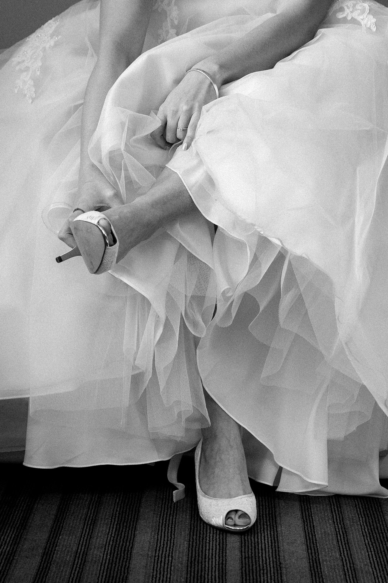 A bride wearing high heels and a wedding dress, sitting on a bench with her legs crossed, adjusting her shoe.