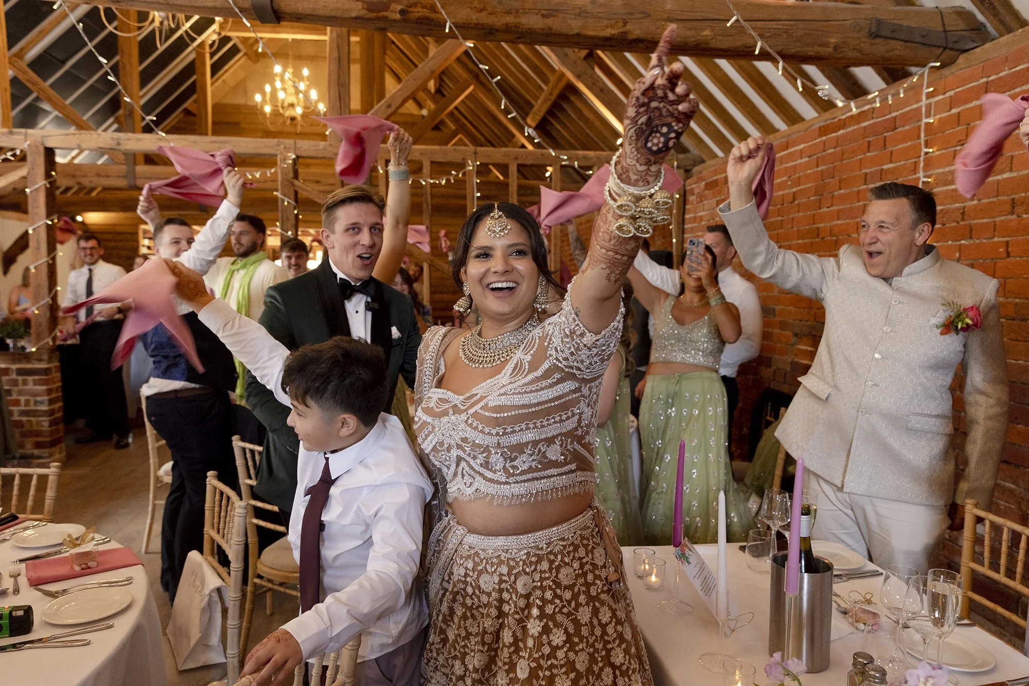 Celebrating at a wedding reception with a woman in traditional Indian attire raising her hand joyfully, surrounded by guests dancing and smiling in a decorated rustic indoor venue.