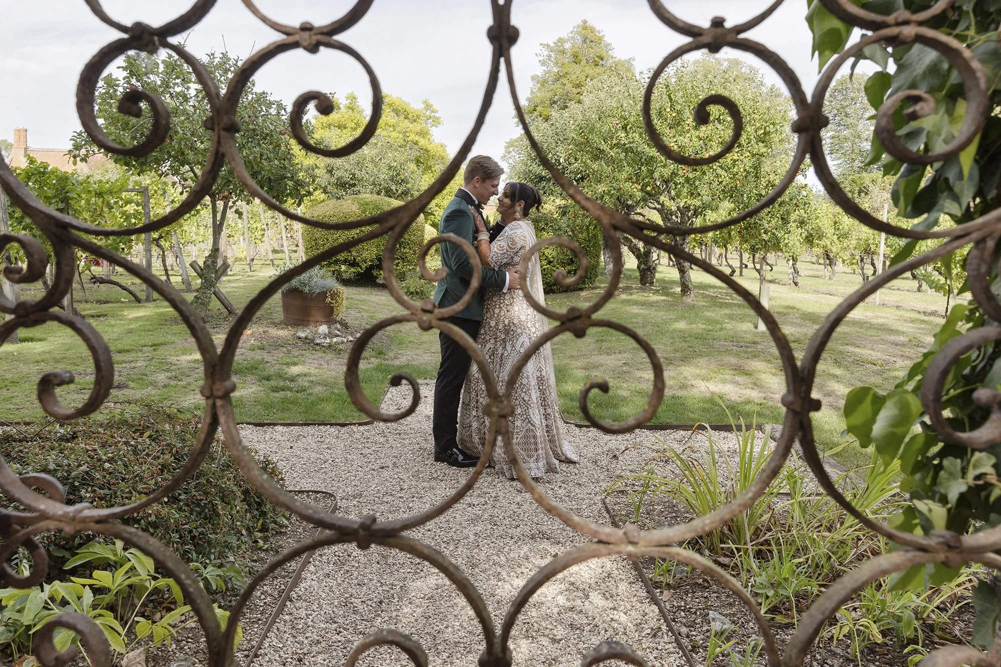 A couple is standing close together inside a garden, seen through a decorative wrought-iron fence, with trees and greenery in the background.