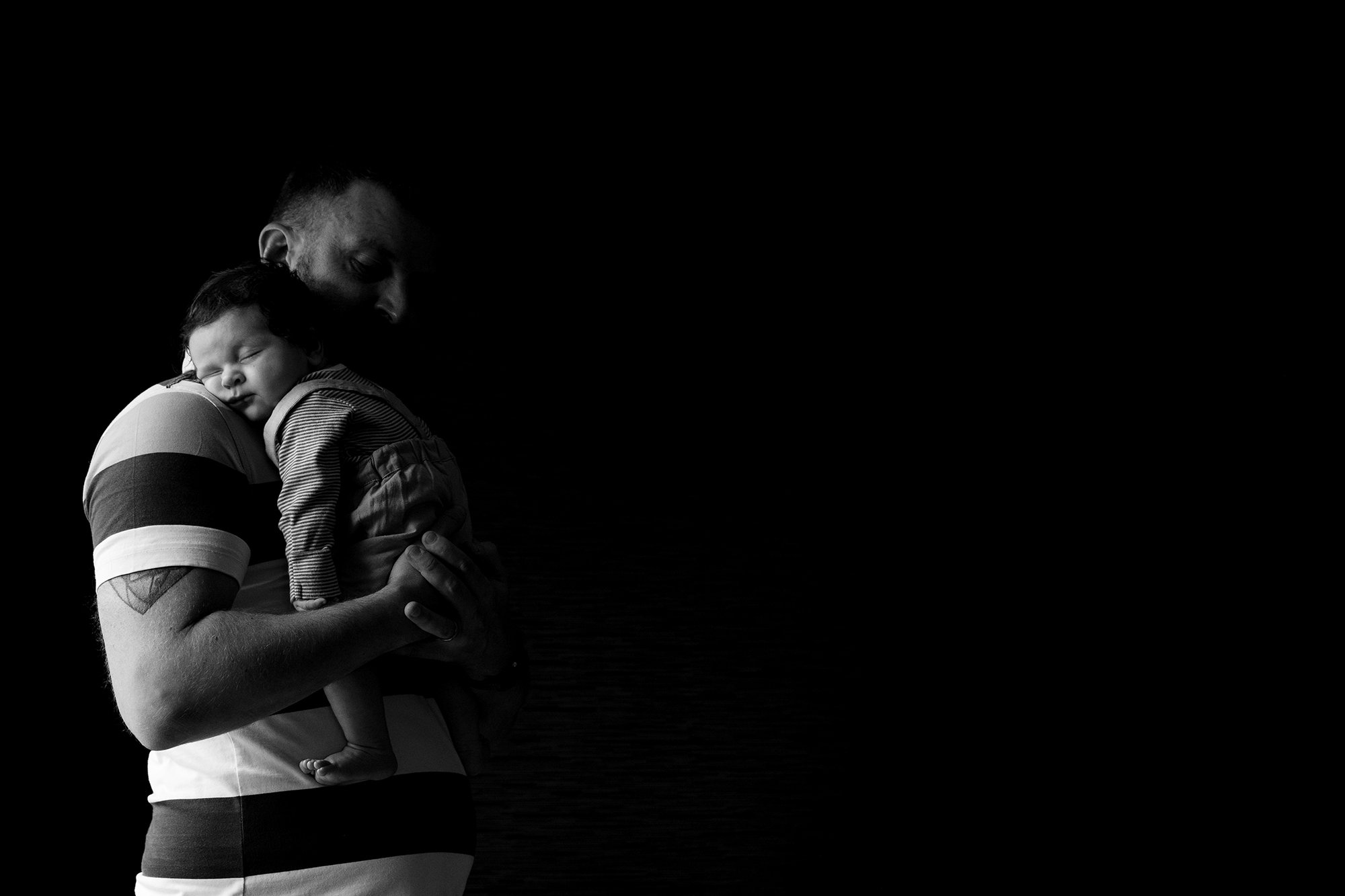 A man is holding a sleeping baby against his shoulder, with a dark background.