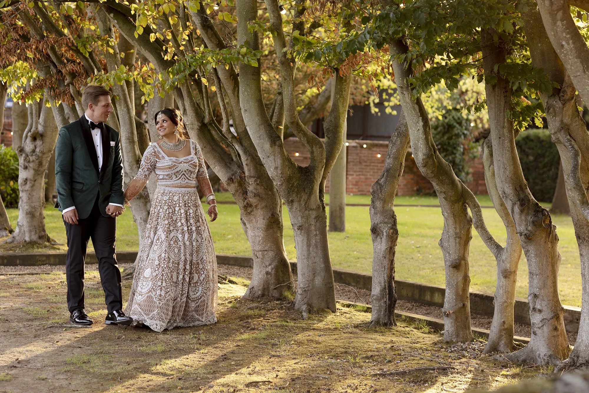 A bride and groom holding hands and walking together outdoors in a park during sunset, surrounded by trees with green leaves.