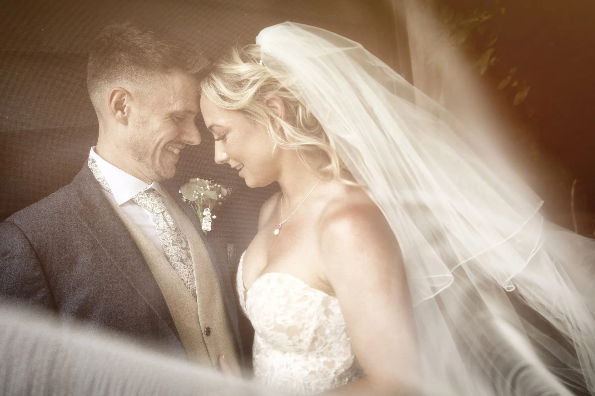 A bride and groom embracing, touching foreheads, with the bride wearing a veil and a lace gown, and the groom in a suit with a tie and boutonnière.