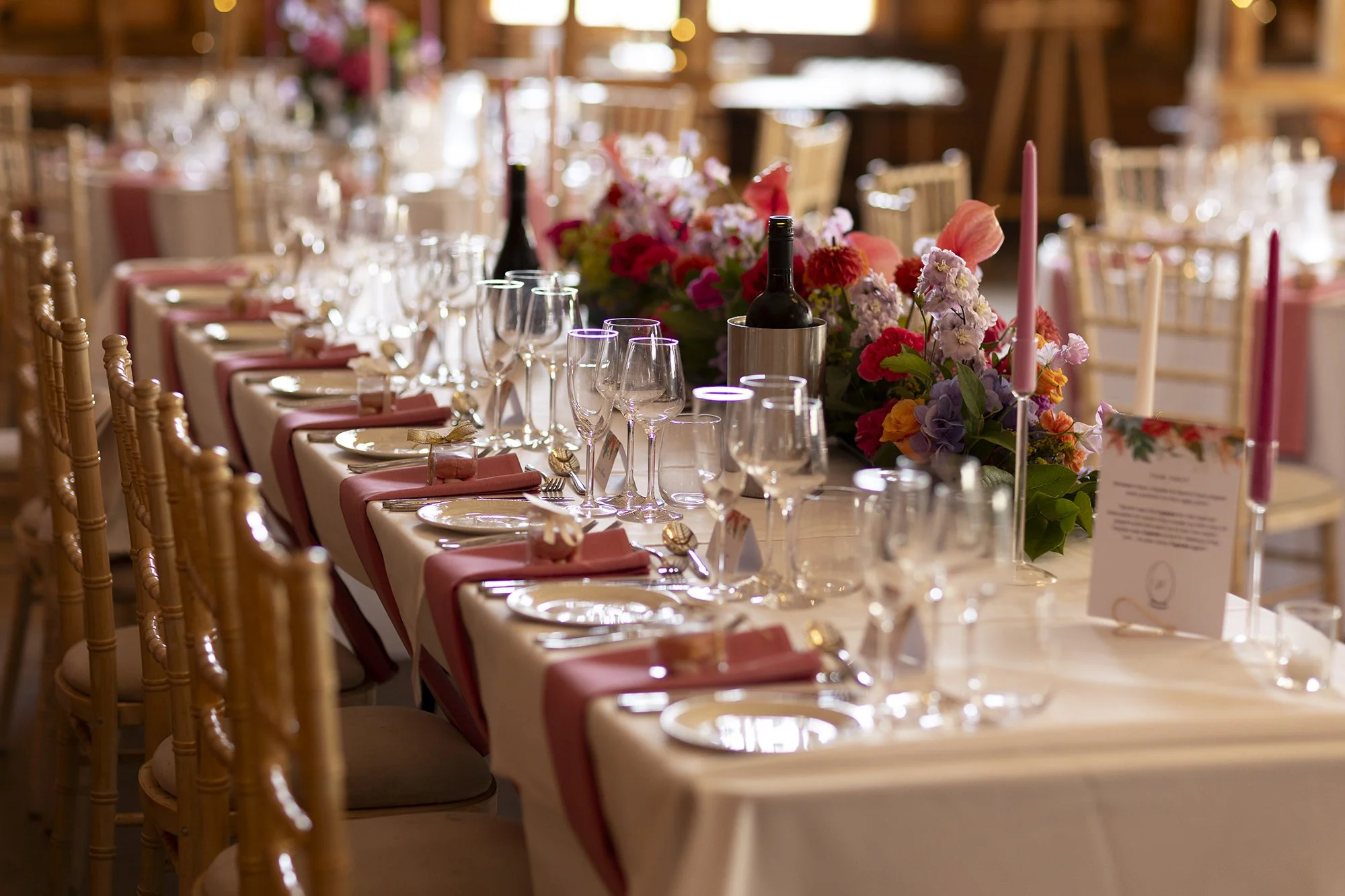A beautifully decorated banquet table set for a wedding reception with pink napkins, white plates, gold utensils, wine glasses, and floral centerpieces with pink, purple, and red flowers.