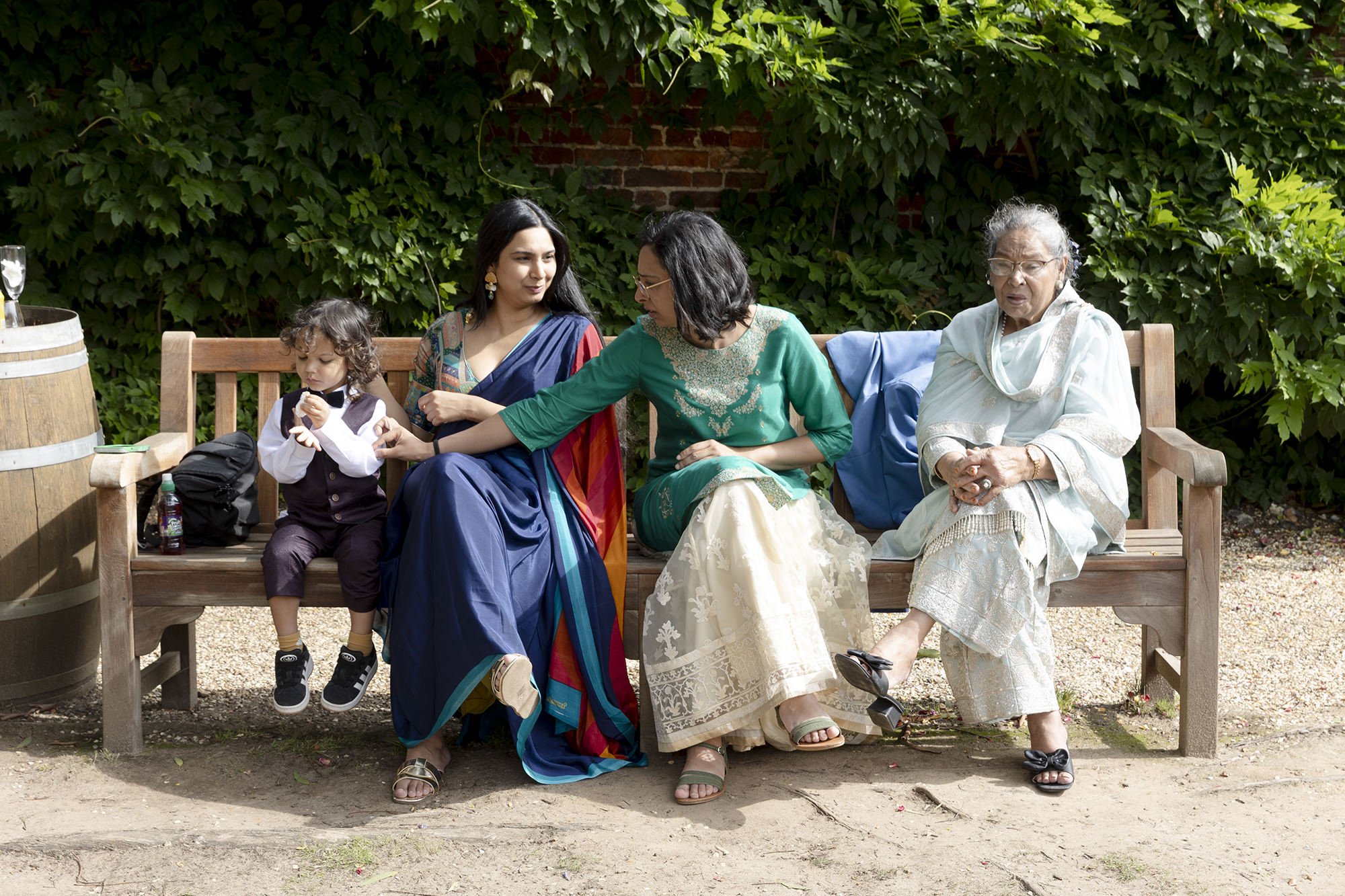Five women and a young boy sitting on a wooden bench outdoors with green foliage behind them, engaged in conversation.