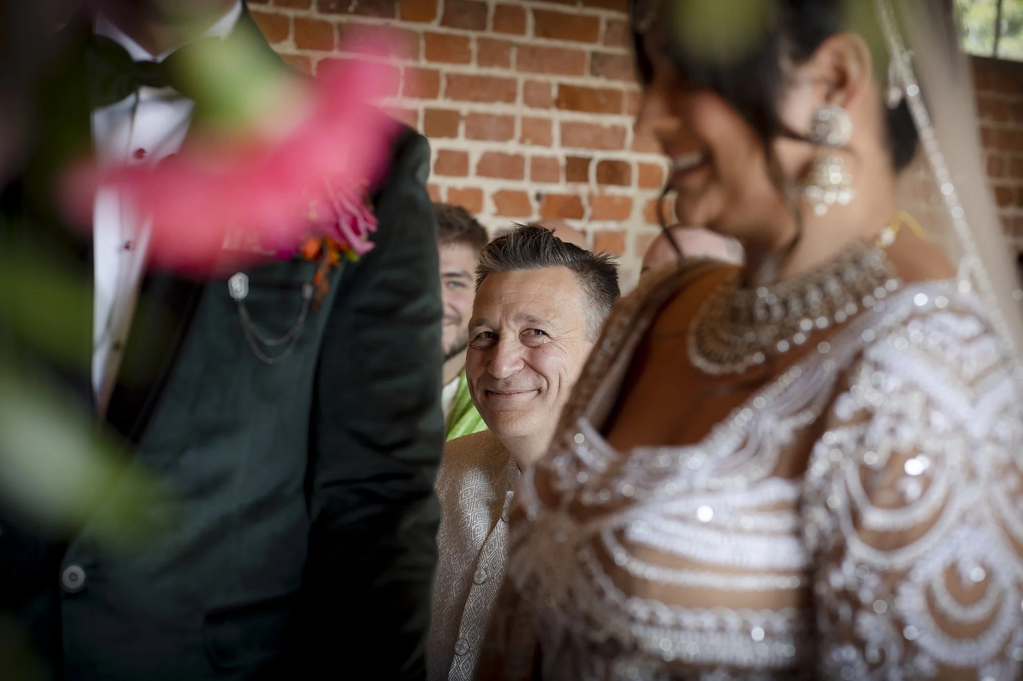 A groom and bride during a wedding ceremony, with the groom smiling and the bride wearing an ornate dress, while another man looks on in the background.