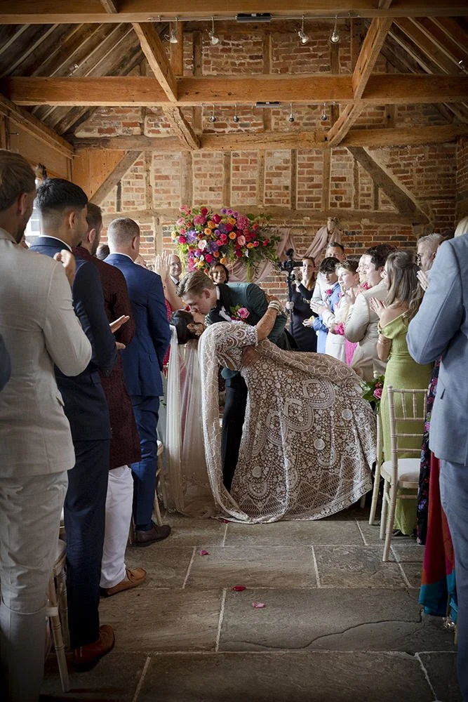 Bride and groom kissing under wedding canopy with flowers in a rustic indoor setting with brick walls, surrounded by guests.