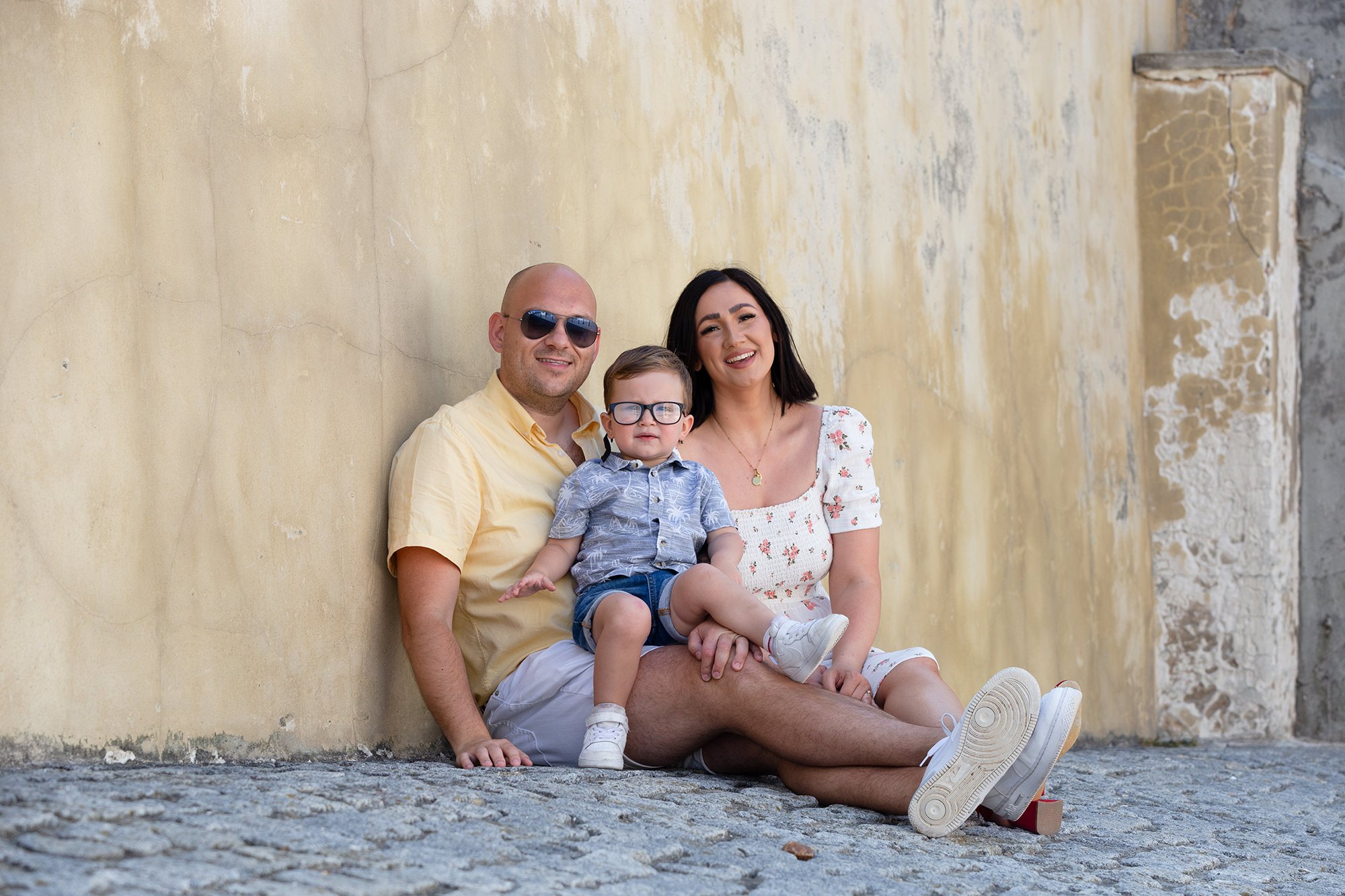 A family of three sitting on cobblestone street against a textured yellow wall, smiling and relaxed. The father wears sunglasses and a yellow shirt, the mother wears a white floral dress, and the young boy wears glasses and a patterned shirt.
