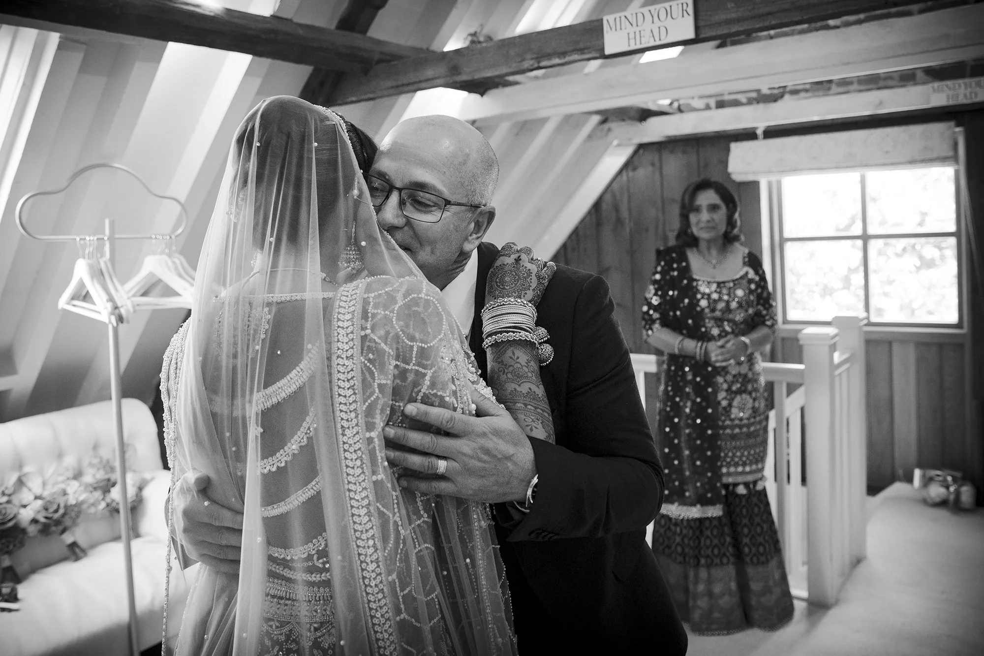A wedding scene with a bride and an older man, possibly her father, embracing in a heartfelt hug inside an attic room with wooden walls. The bride is wearing a traditional dress with a veil, and the man is dressed in a suit and glasses. A woman in tr