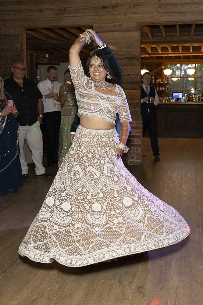 A woman in a traditional Indian outfit dancing at a celebration or wedding. She is wearing an ornate cream-colored lehenga with detailed embroidery. People are watching her in the background inside a rustic wooden venue.