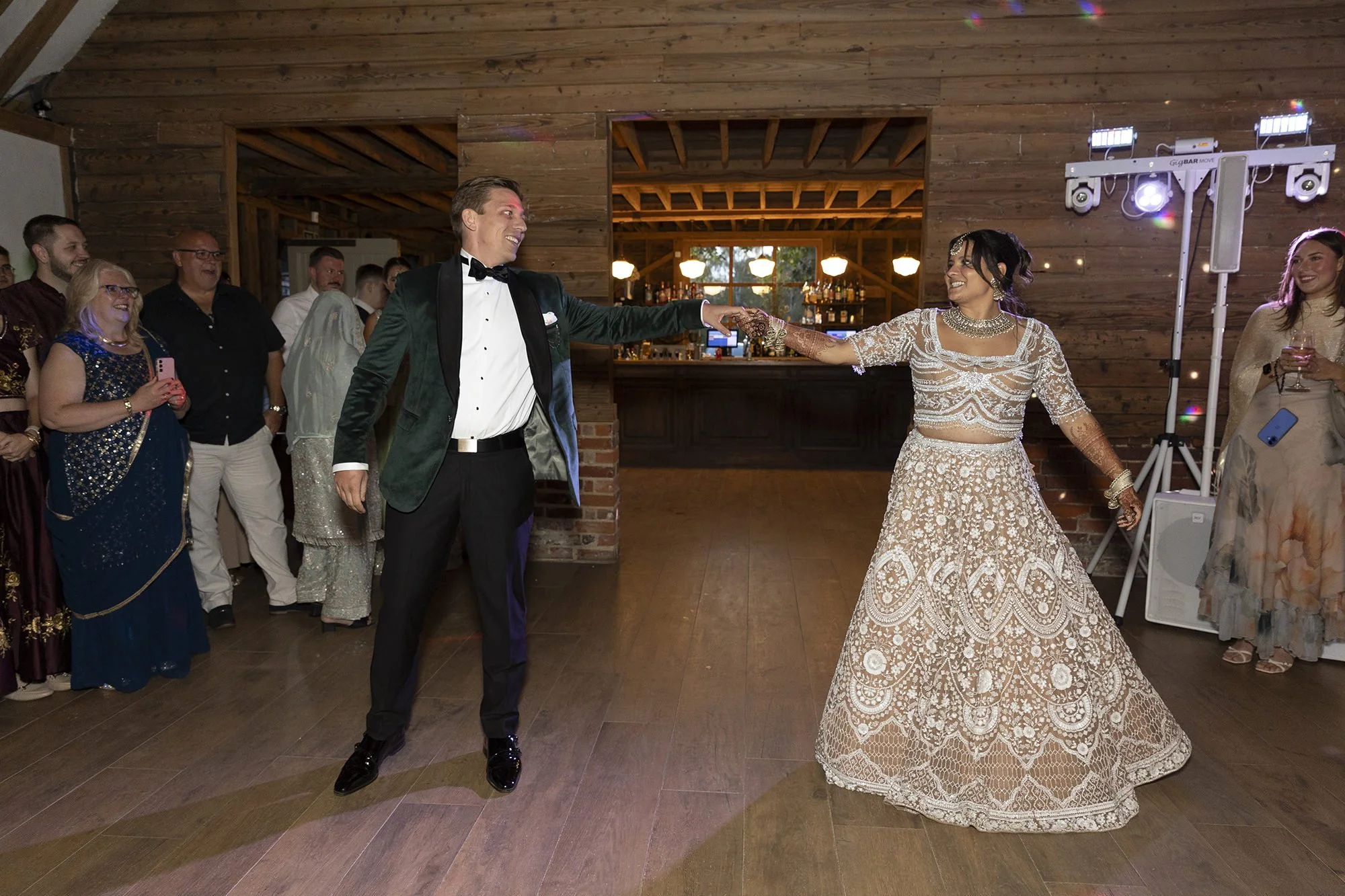 A bride and groom dancing at their wedding reception with friends and family watching and smiling in the background. The bride is wearing a cream-colored, intricate traditional dress, and the groom is in a dark green velvet blazer with a black bow ti