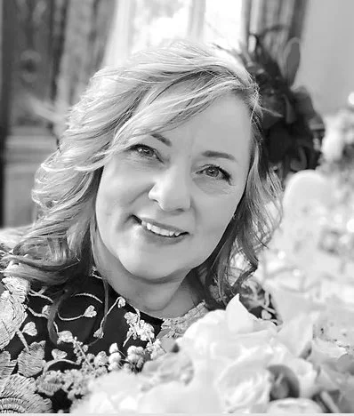 A woman with wavy hair smiling at the camera, surrounded by flowers and plants in what appears to be a greenhouse or plant shop.