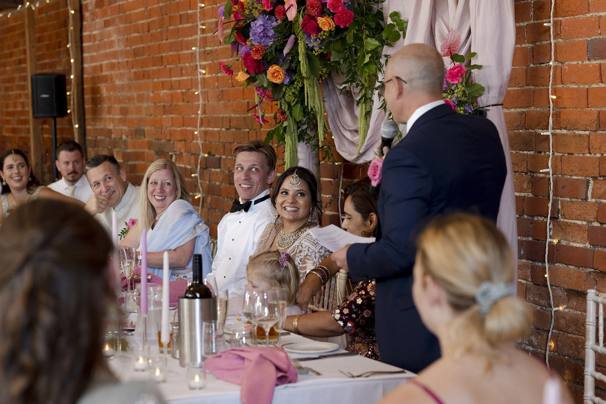 A wedding reception with a groom, bride, and family members sitting at a decorated table, listening to a man giving a speech with a microphone. The background features a brick wall and floral decorations.