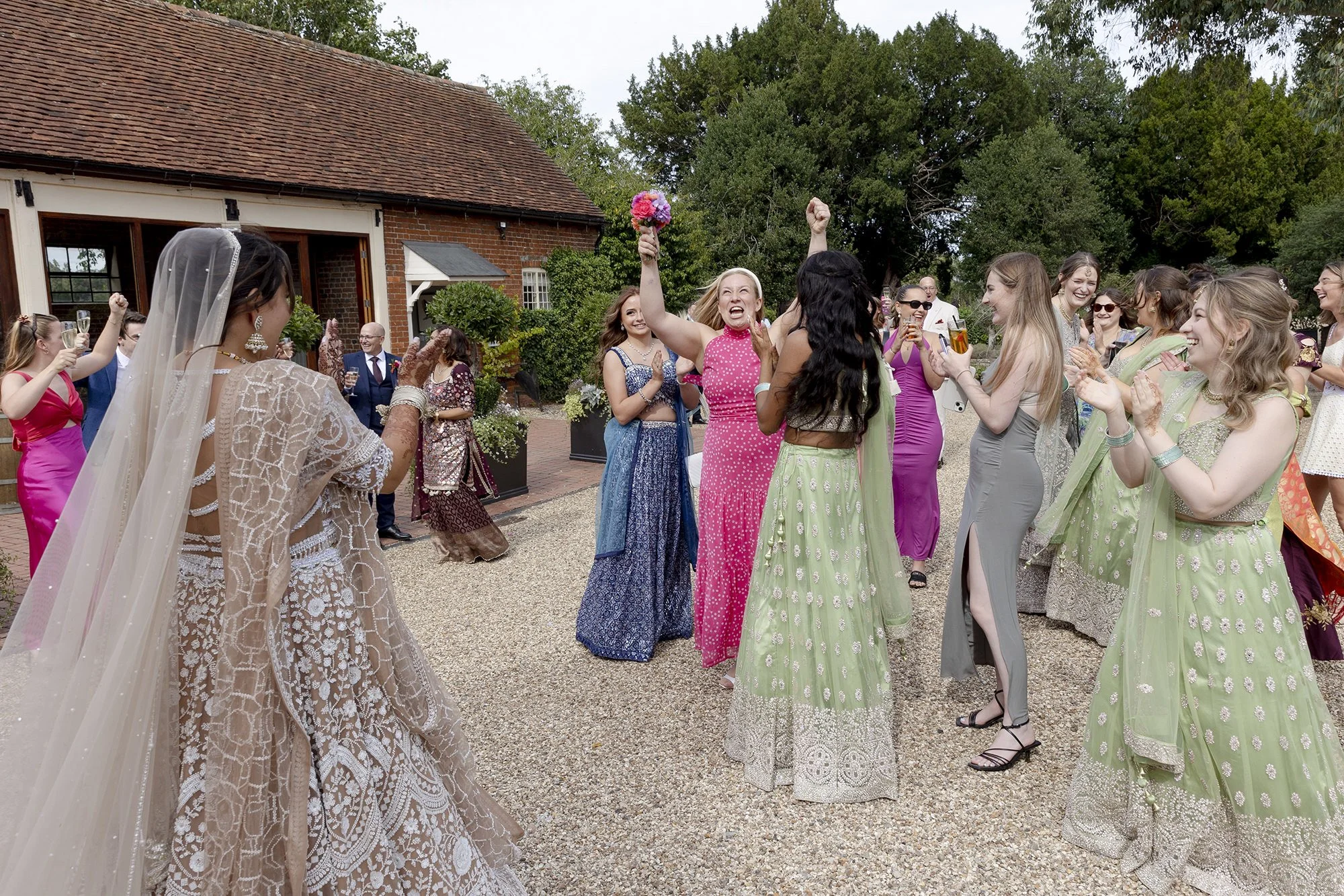 People celebrating outdoors at a wedding, with women in colorful dresses, some wearing traditional South Asian attire, smiling and clapping, with one woman holding a bouquet and another with her fist raised in triumph.