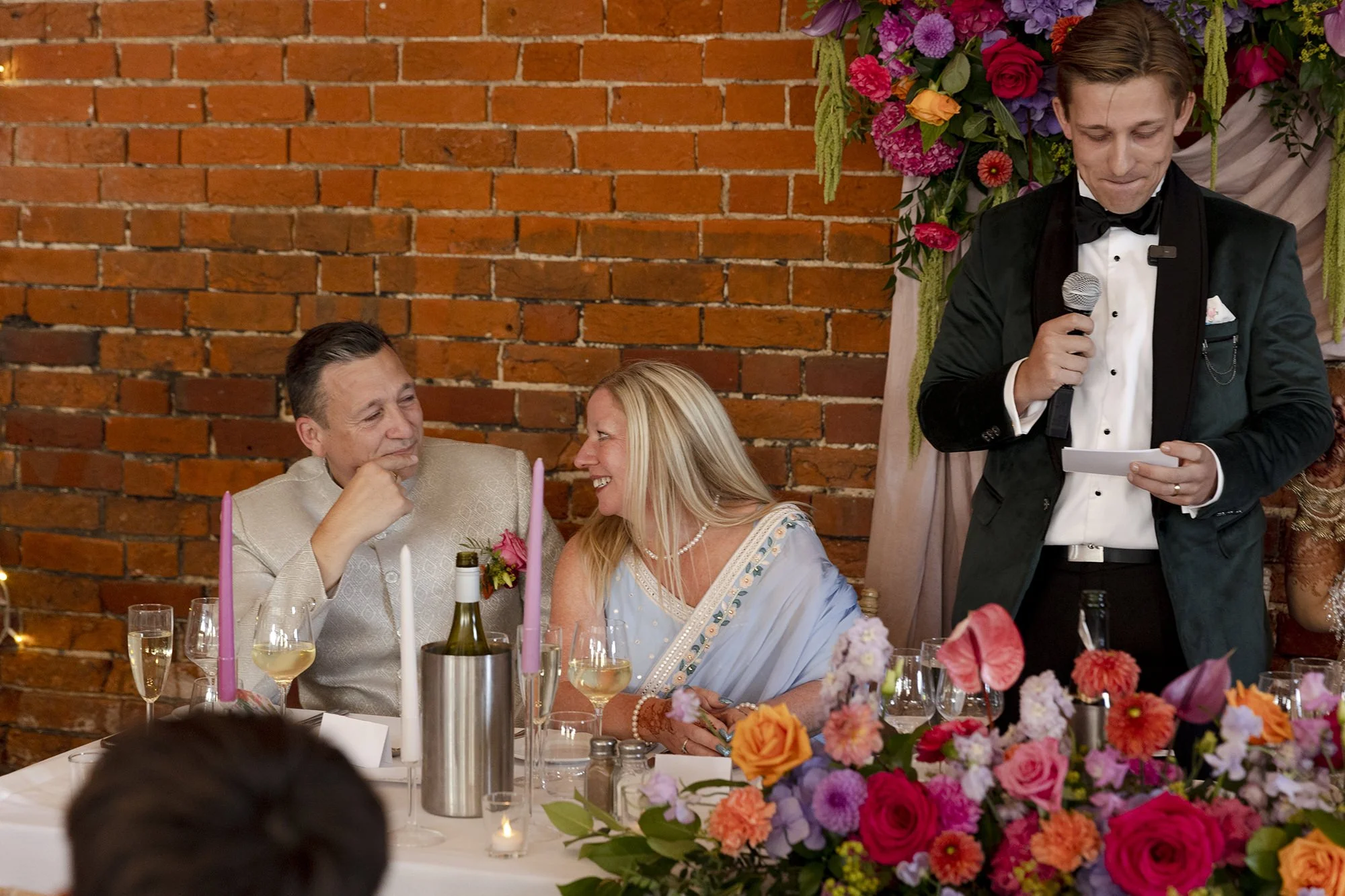 People sitting at a decorated table with flowers and candles at a wedding reception, with a man giving a speech while holding a microphone and notes.