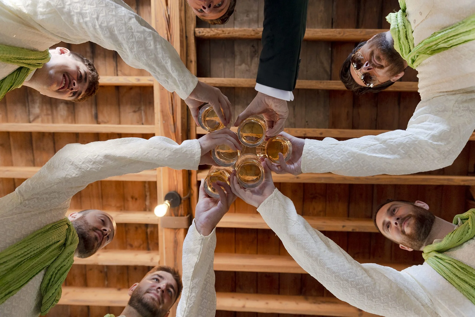A group of seven people in traditional Indian attire toasting with glasses of whiskey inside a wooden structure.