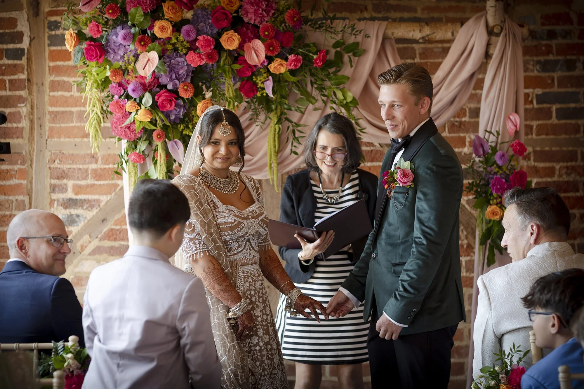 A bride and groom holding hands during their wedding ceremony, standing in front of a floral arch, with a officiant reading from a book, surrounded by seated guests in a rustic brick venue.