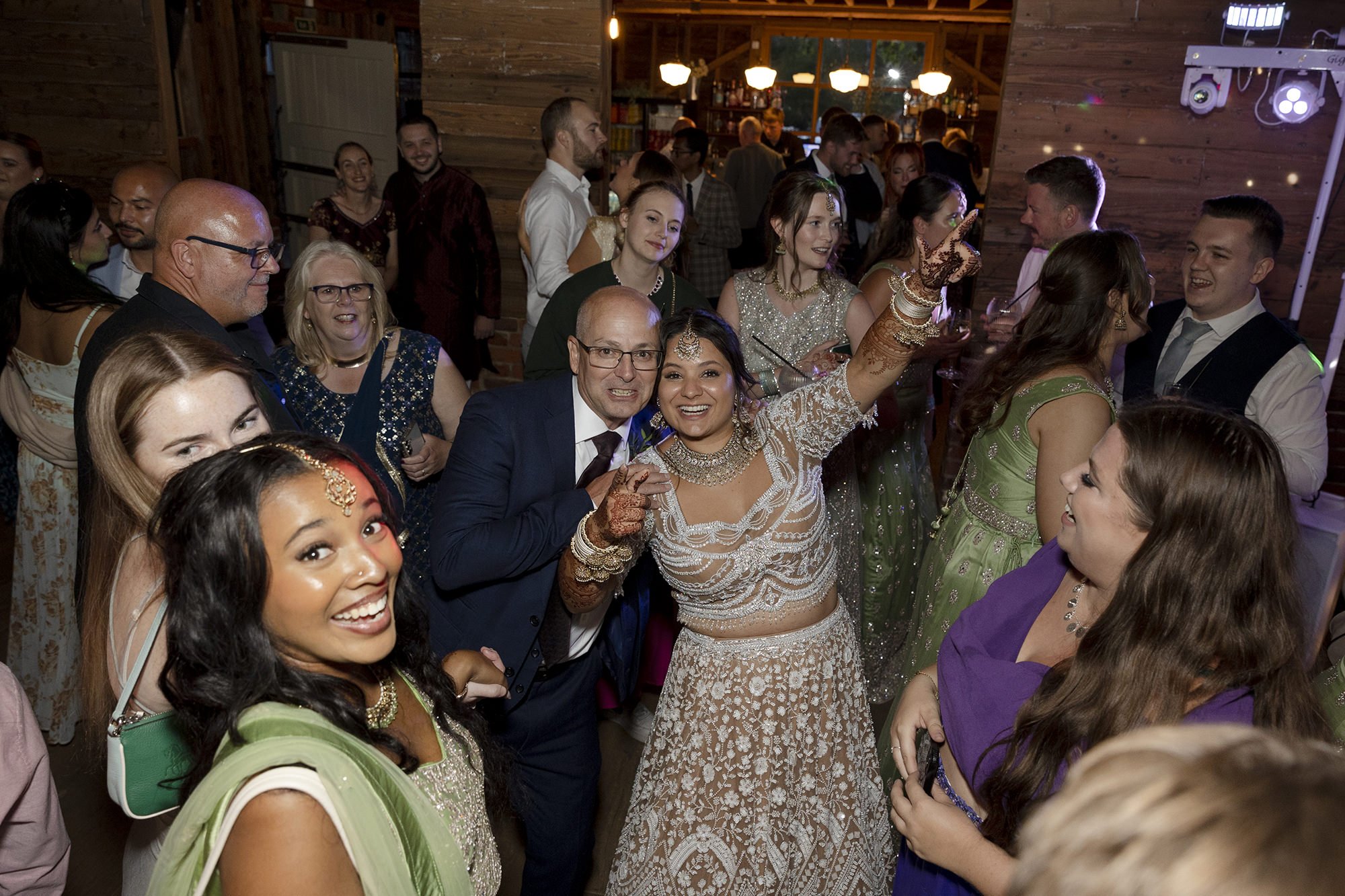 People celebrating at a wedding reception indoors, dressed in colorful traditional Indian attire, with illuminated wood-paneled walls and a bar in the background.