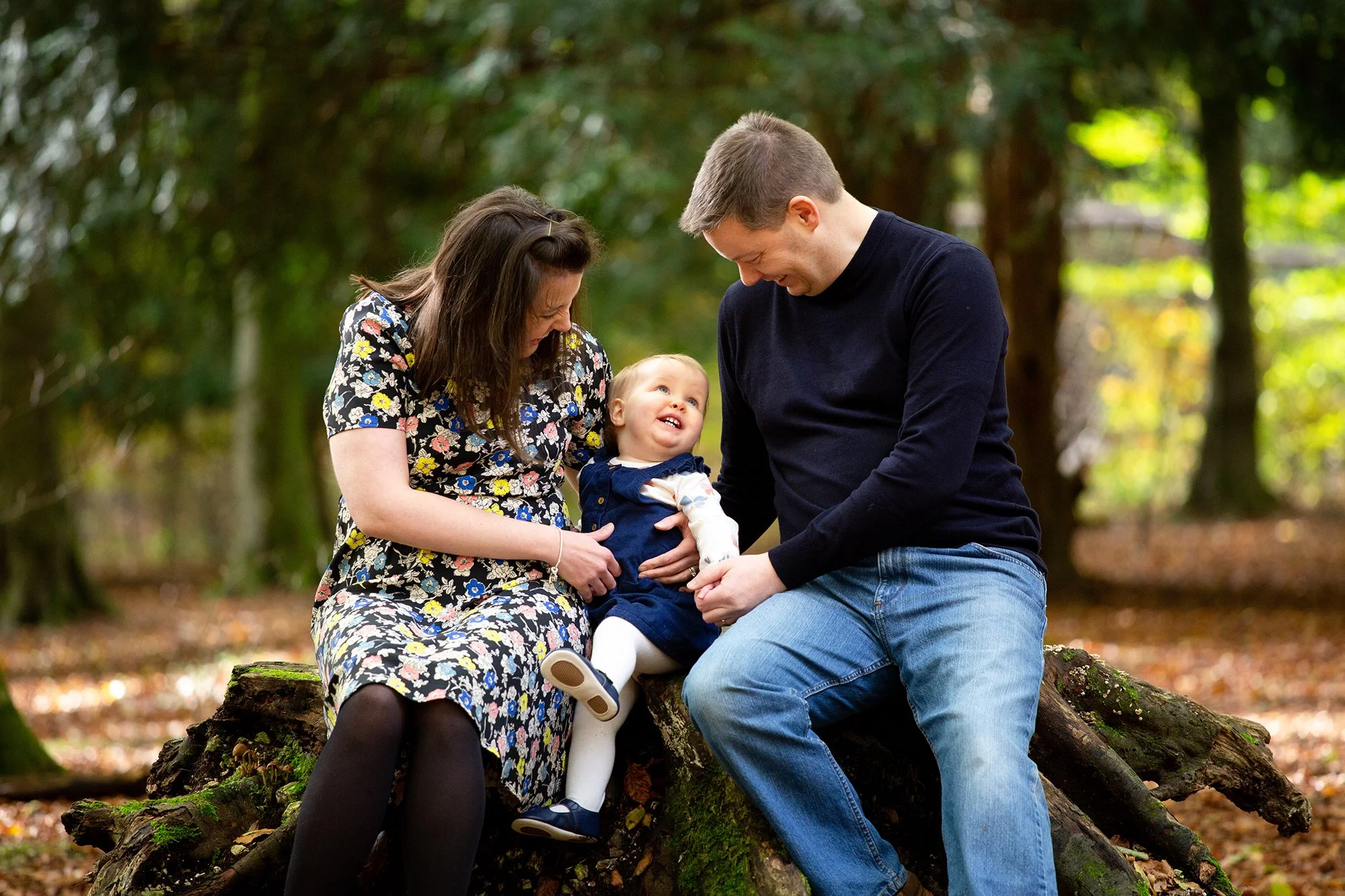 A family of three sitting together on a fallen tree in a forest, smiling and enjoying each other's company.