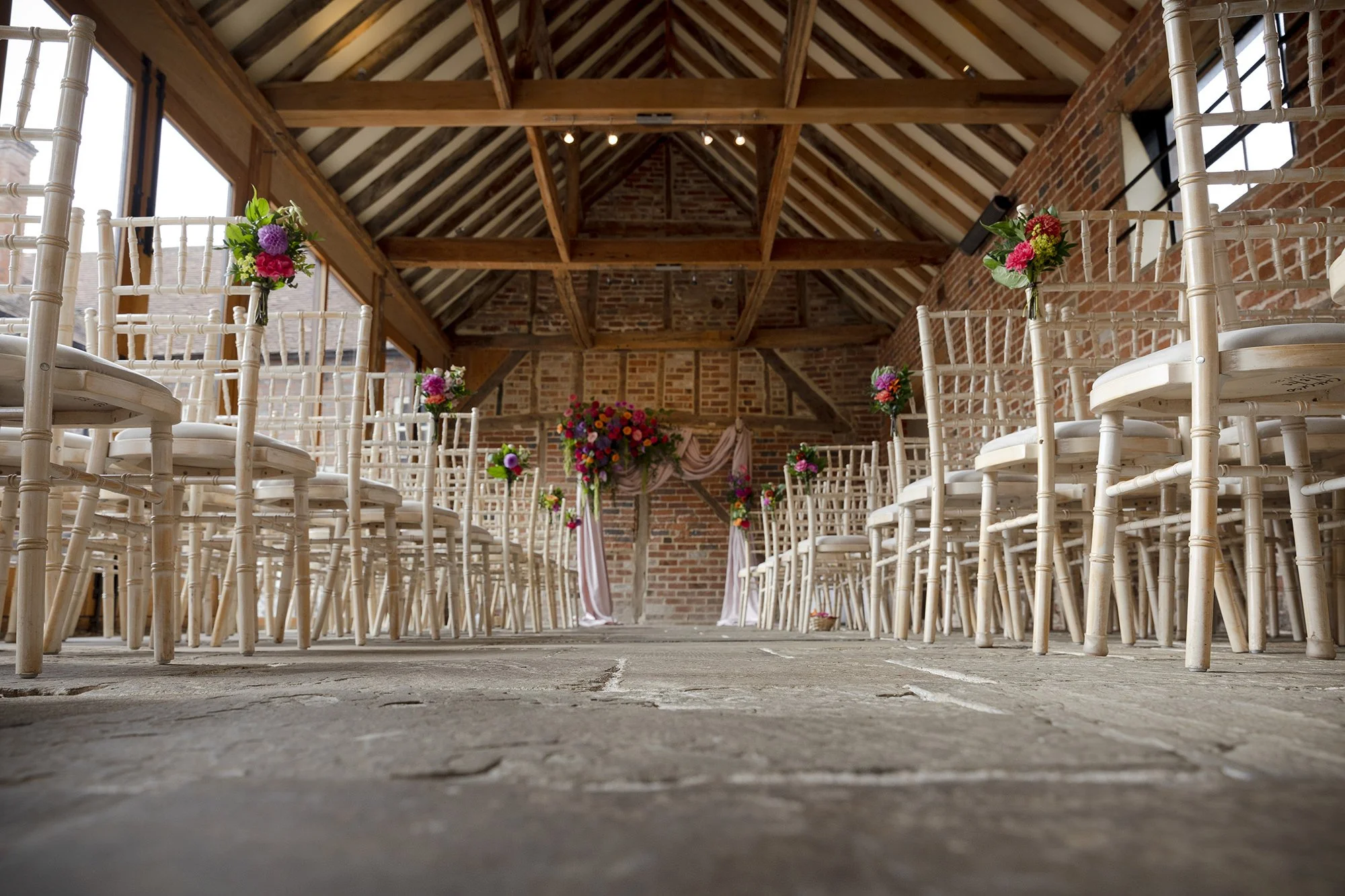 Wedding ceremony setup in a rustic barn with white chairs decorated with colorful floral arrangements along the aisle leading to a floral arch at the front.