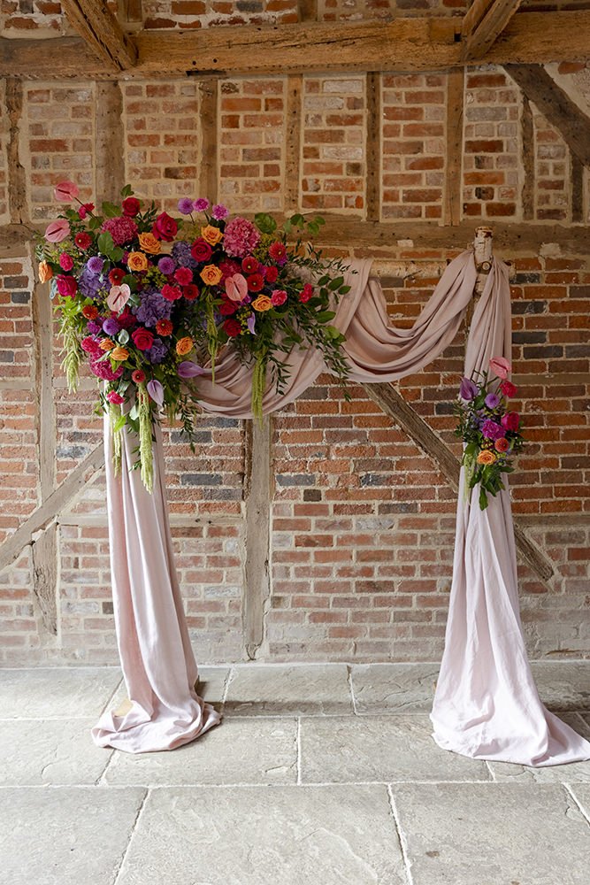 Decorated wedding arch with pink drapery and a large floral arrangement featuring red, pink, orange, purple flowers, and greenery, against a brick wall.
