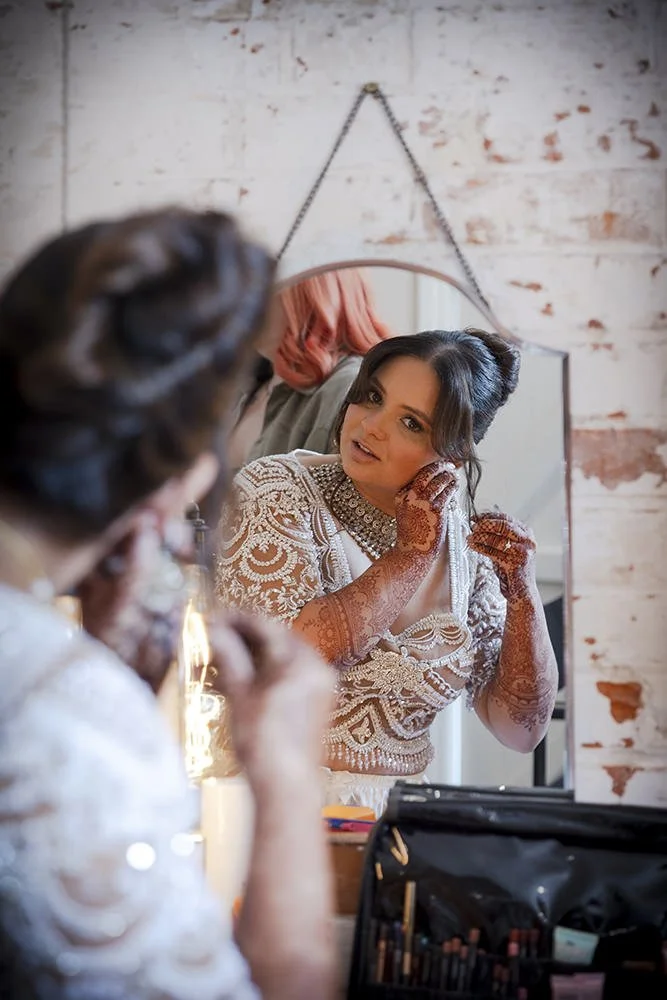 A woman with intricate henna tattoos on her hands and arms, wearing a lace and embroidered dress, is looking at herself in a mirror as she adjusts her earring, with a makeup kit and a person with pink hair reflected in the background.