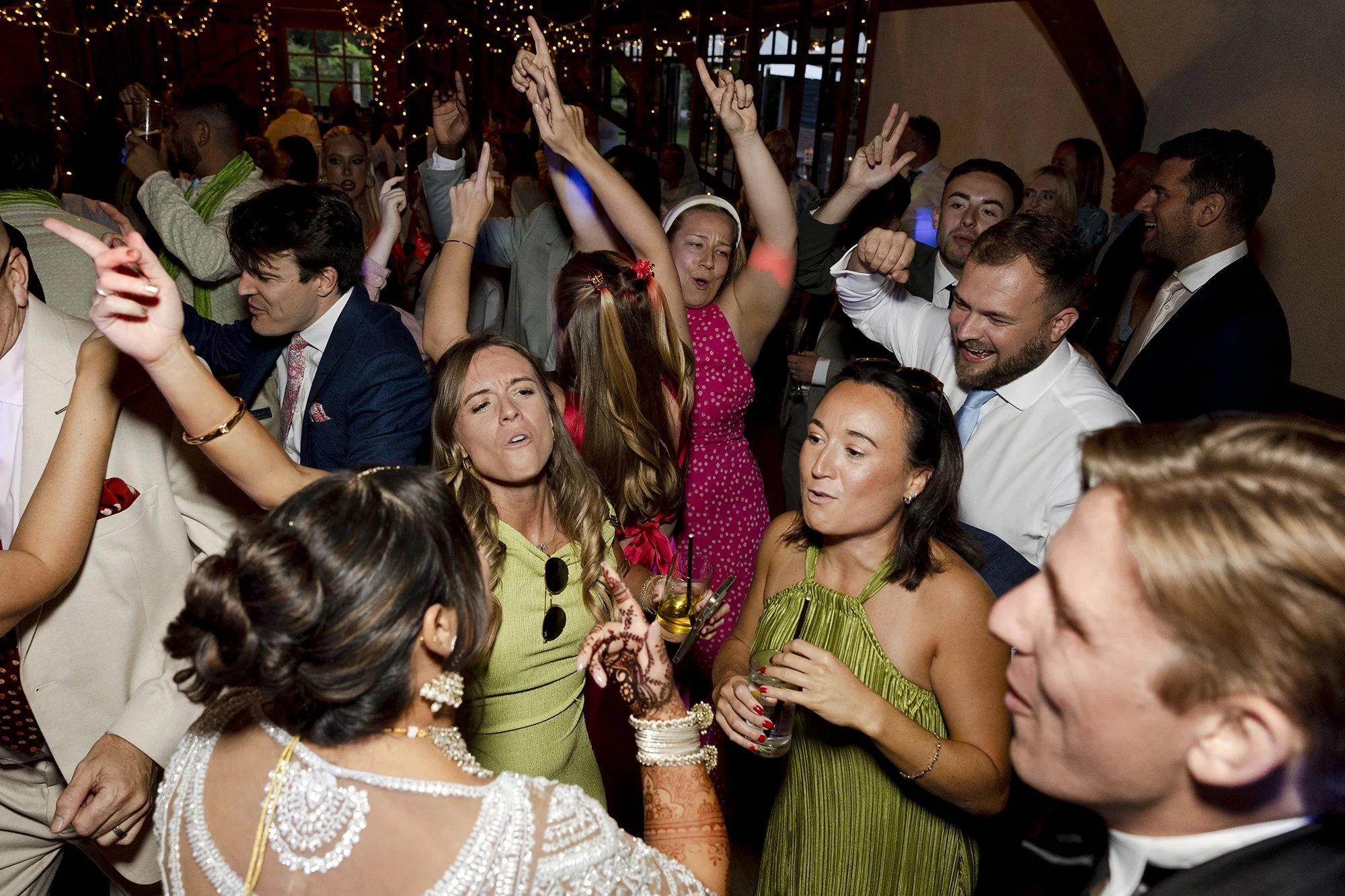 People dancing and socializing at a lively event or party, with some raising their hands and others holding drinks, in a decorated indoor venue.