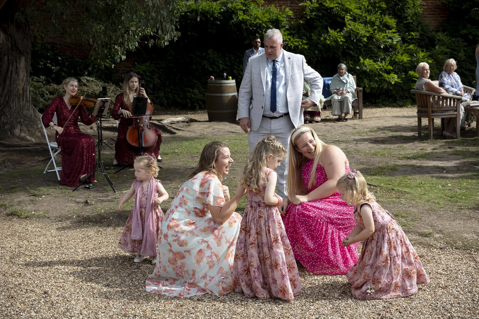 Celebration scene outdoors with children in floral dresses dancing with an adult woman, while a man in a light suit observes. Musicians with violins and a cello play in the background, and other older adults sit on benches.