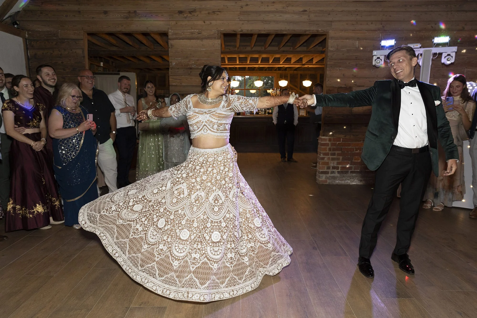 A bride and groom dance together at their wedding reception, with guests watching and taking photos in a rustic indoor venue.