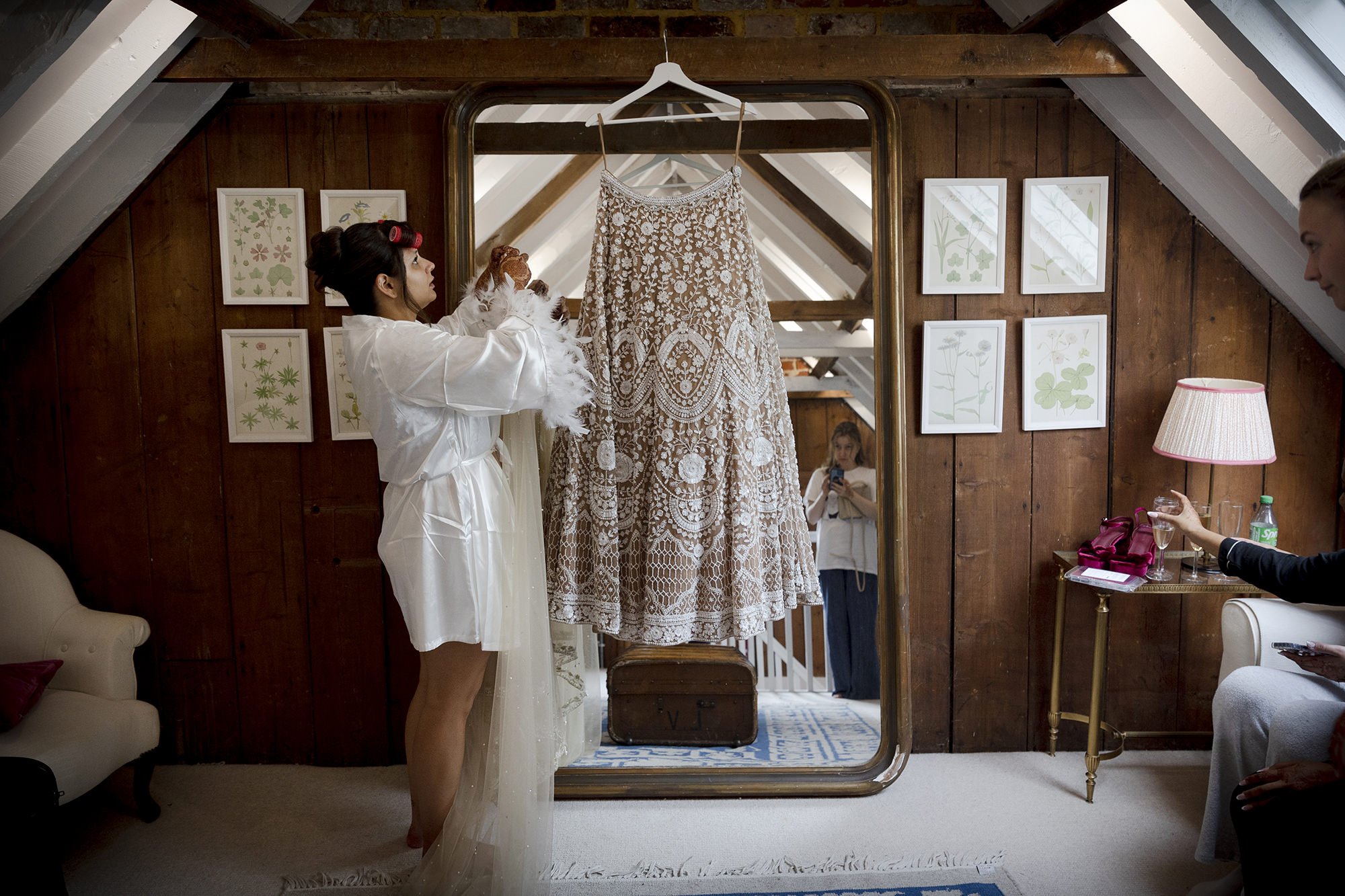 Woman in white robe with curlers in hair examining an ornate lace wedding dress hanging on a mirror in a cozy room.