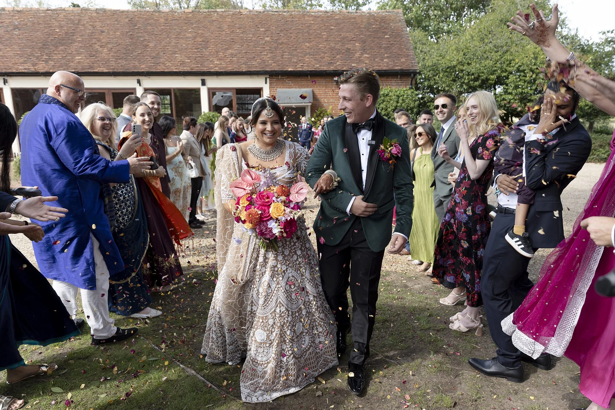 Bride and groom walking arm-in-arm through a crowd of guests at an outdoor wedding, with confetti falling around them.