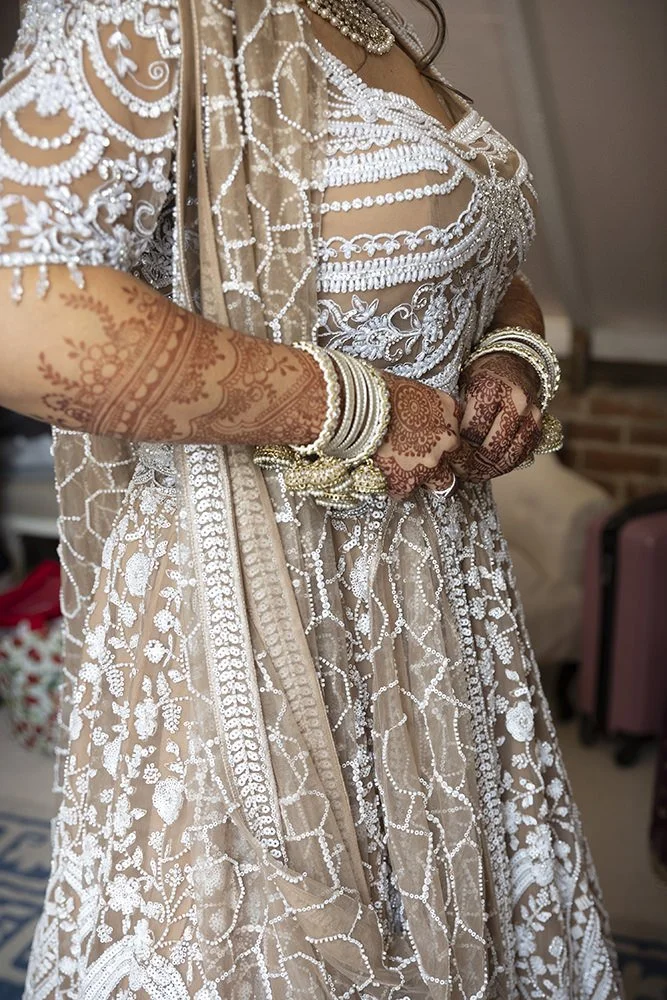 A woman wearing traditional Indian bridal attire with intricate white embroidery and jewelry, including bangles and henna designs on her hands and arms.