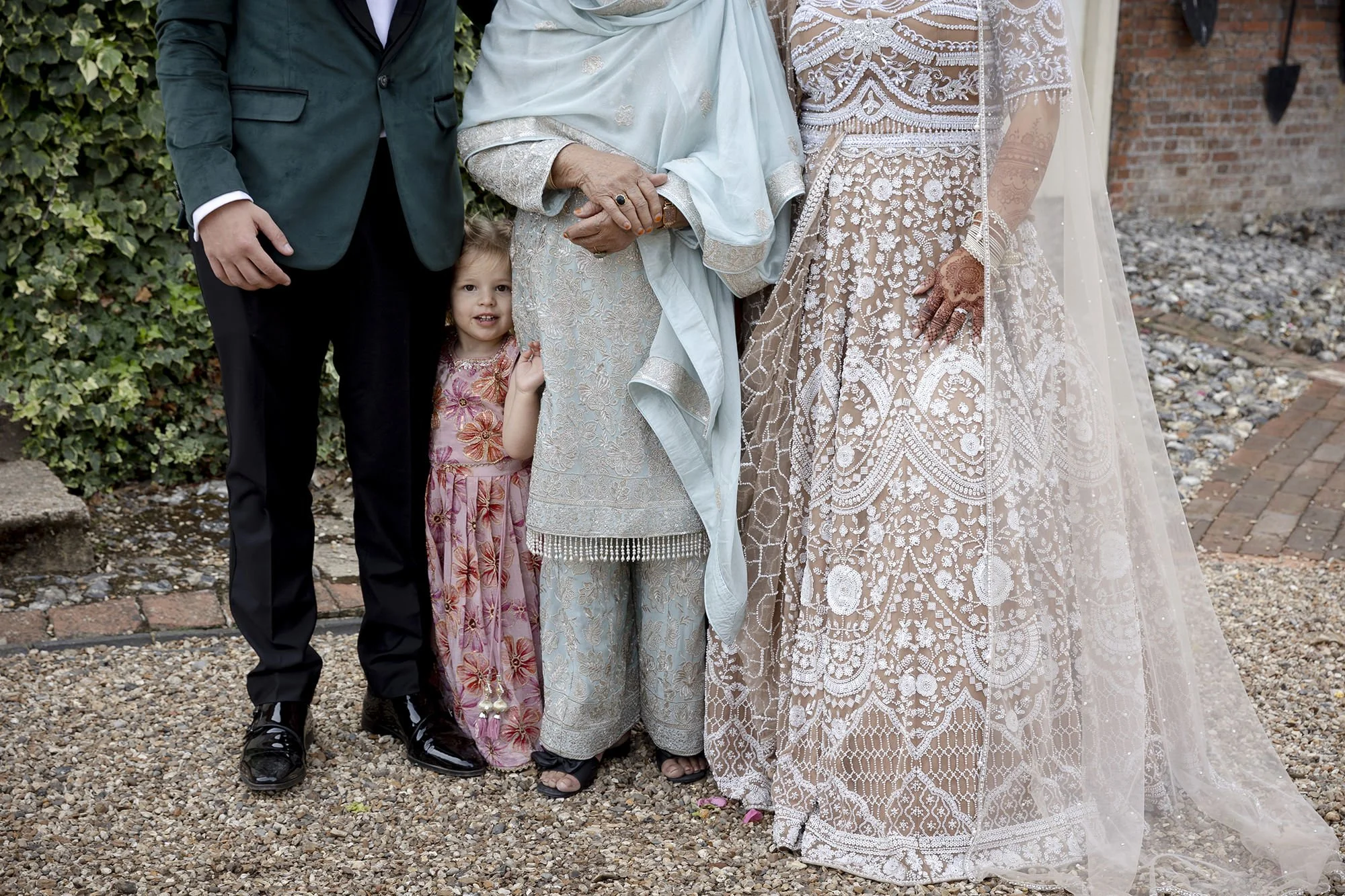 People dressed in formal attire, with a little girl in a floral dress standing between adults in elegant clothing, including a woman in a lace gown, outdoors on gravel ground near a brick wall and greenery.