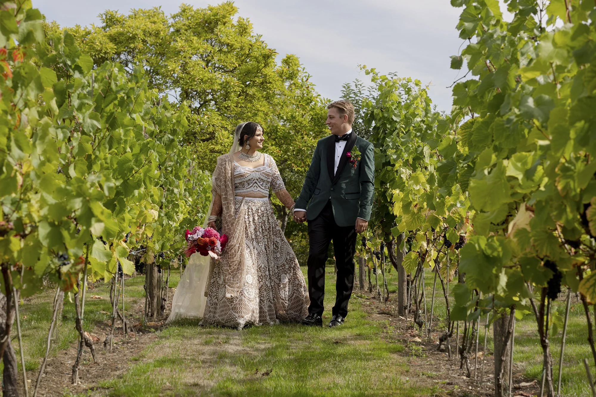 A bride and groom in formal wedding attire holding hands and walking through a vineyard, smiling at each other.