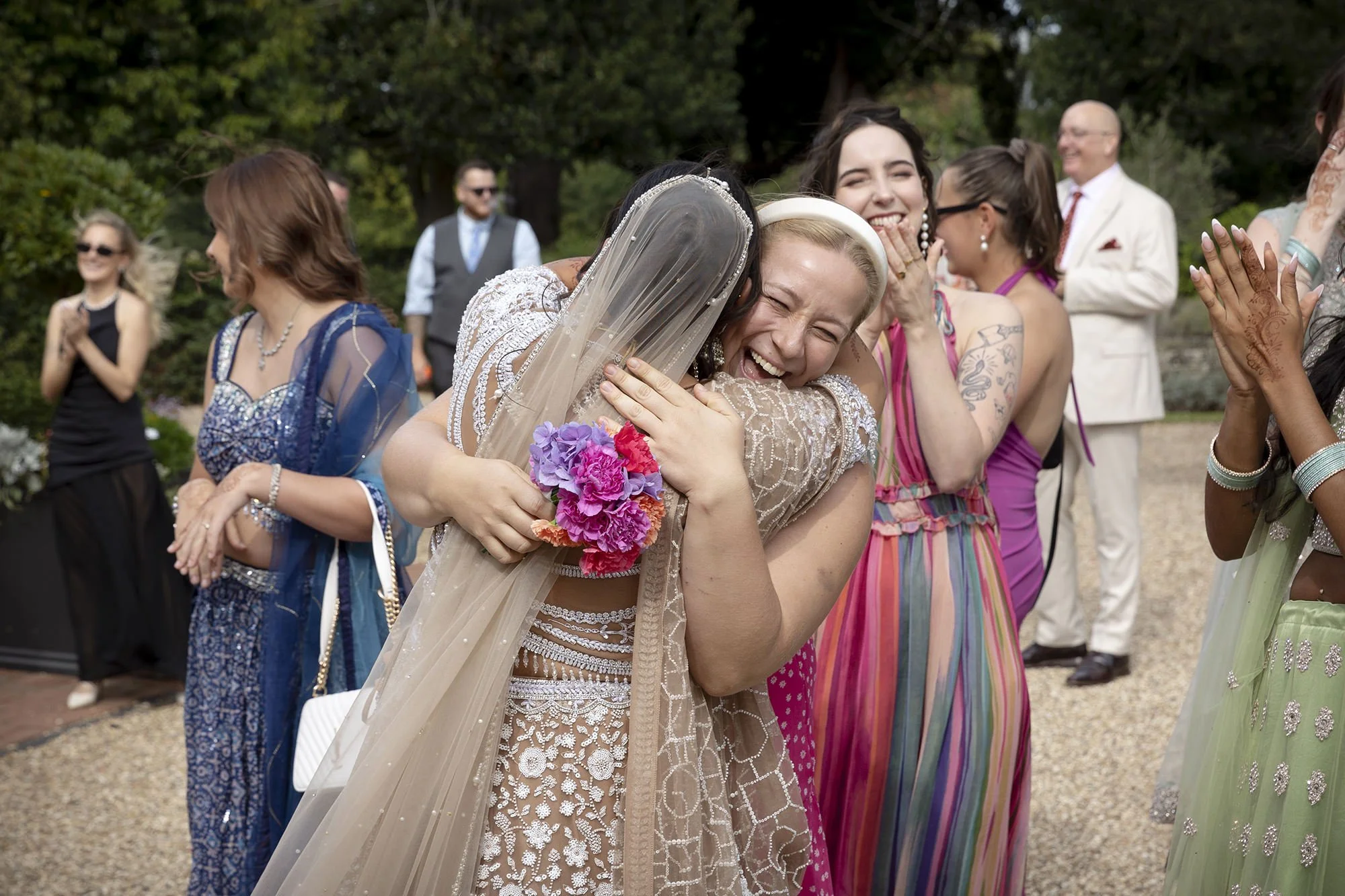 Two women hugging, smiling at a wedding celebration, surrounded by other happy guests outdoors.