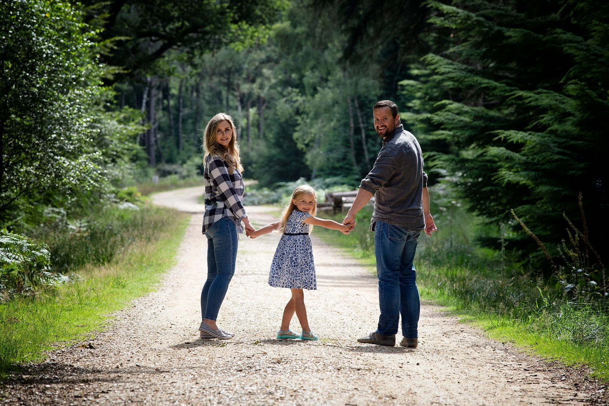 A family of three, a woman, a man, and a young girl, walking hand in hand down a dirt path through a wooded area, smiling at the camera on a sunny day.