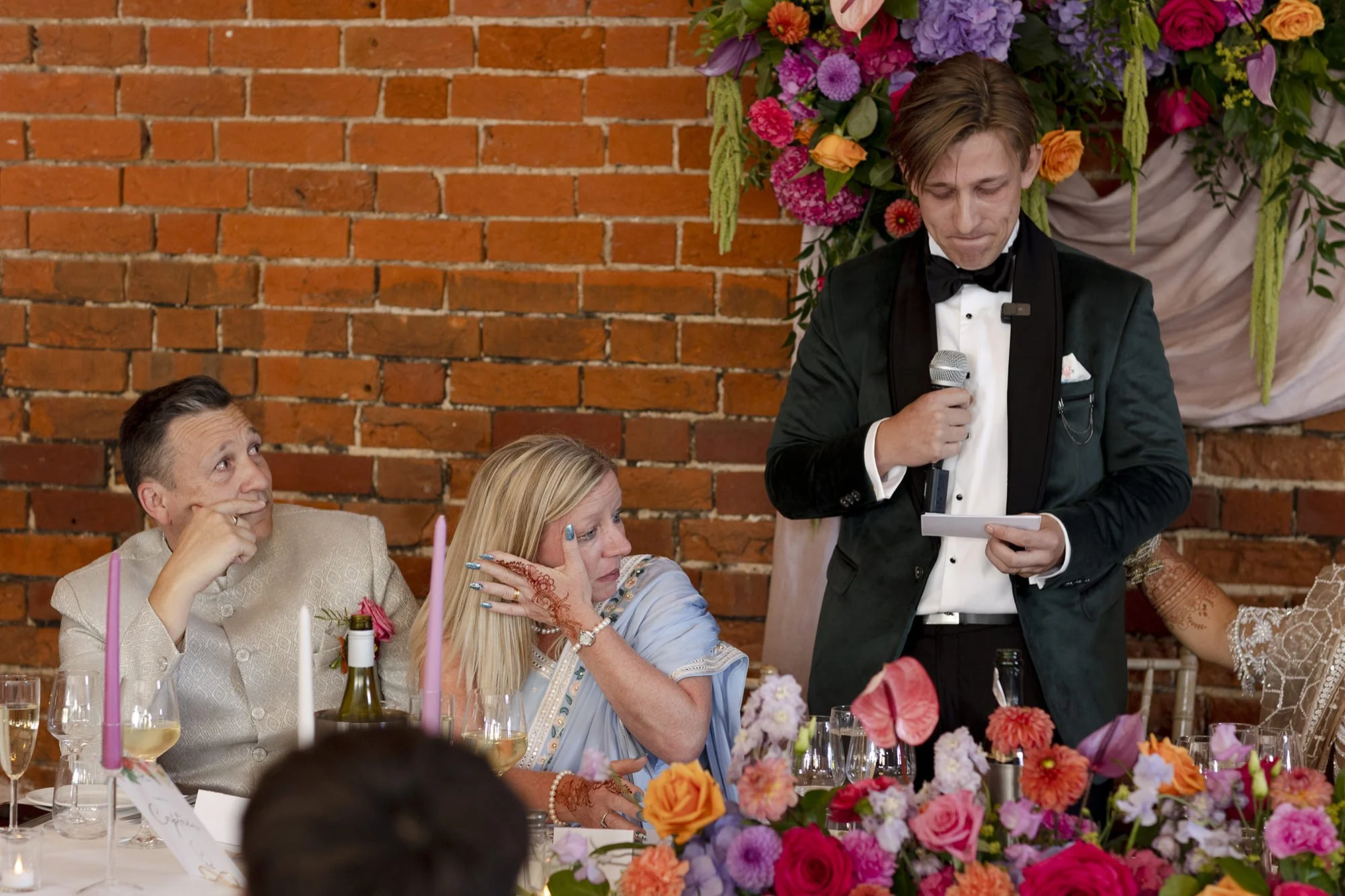 A man in a tuxedo giving a speech at a wedding reception, standing with a microphone in one hand and a card in the other. Two women seated at a table, one old and upset with her hand on her cheek, the other older and thinking with her hand on her fac