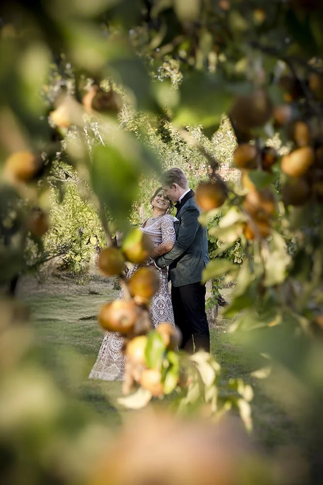 A couple dressed in formal attire standing close together and smiling amid apple trees in an orchard.
