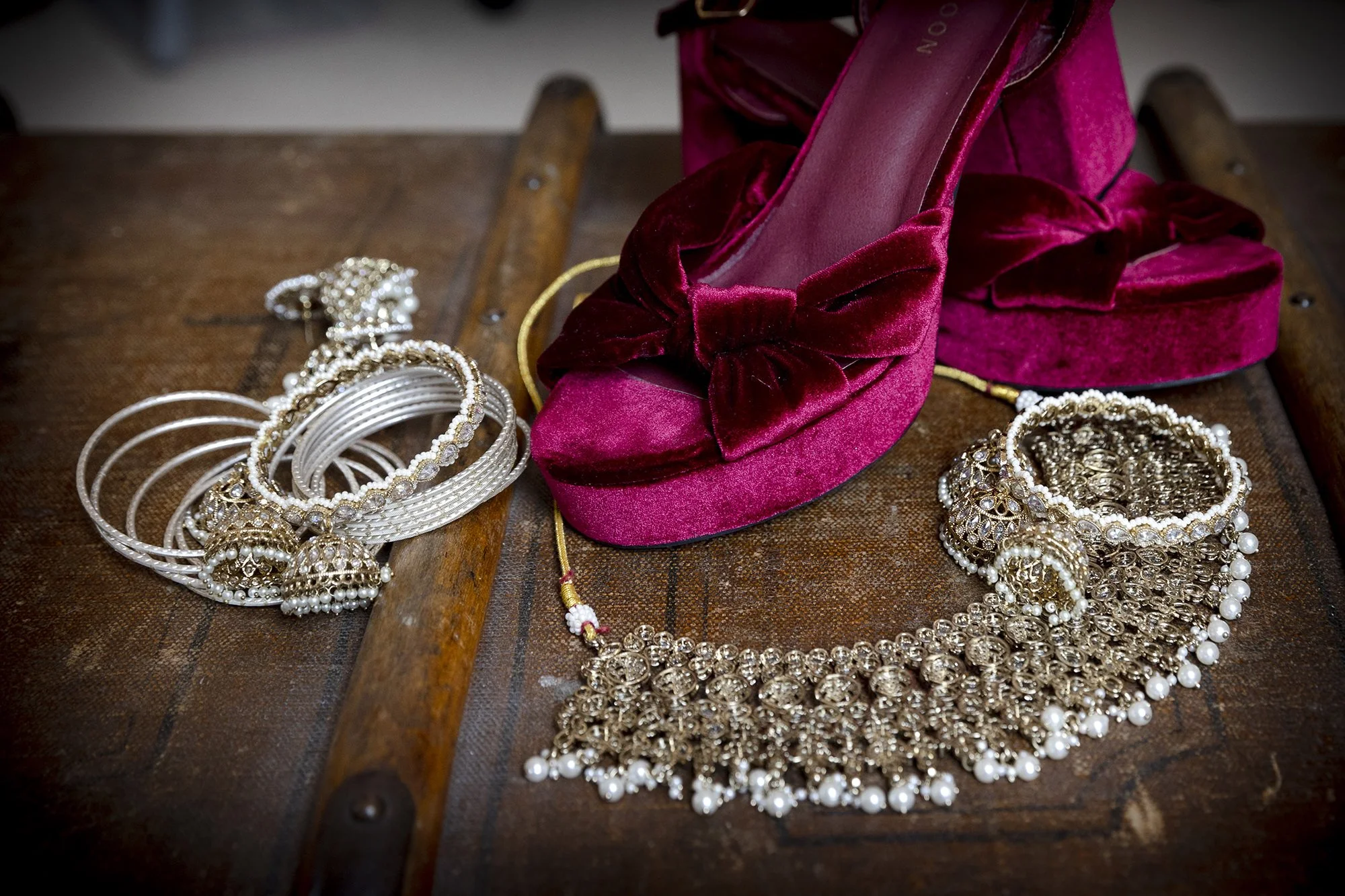 A pair of pink velvet high-heeled shoes with bows, a collection of silver and pearl jewelry including bangles, necklaces, and earrings, all arranged on a wooden surface with a rustic appearance.